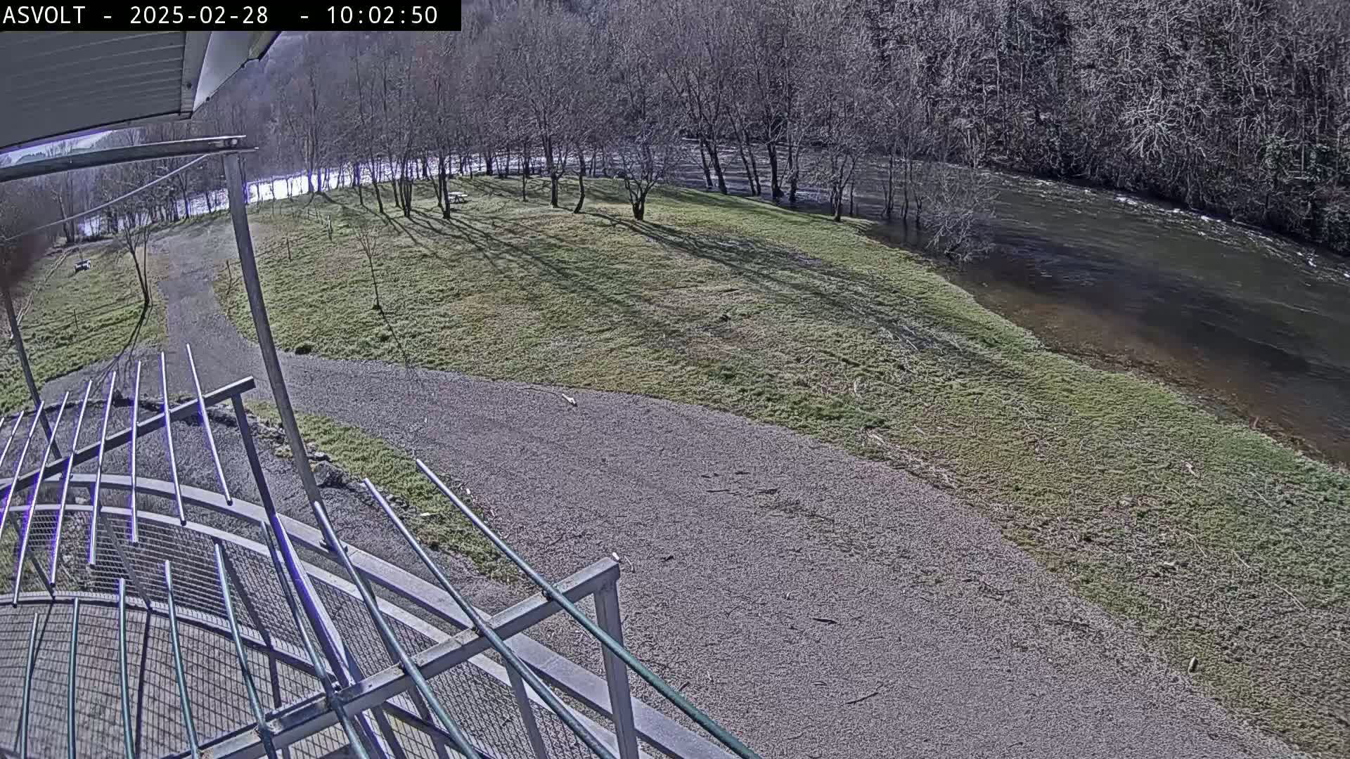 A gravel path leads to a grassy area beside a calm river under a sunny sky, with bare trees lining the riverbank.