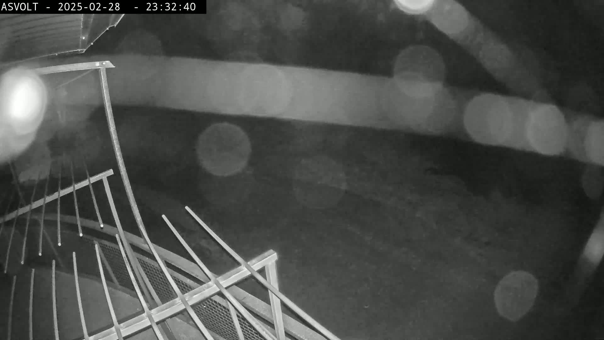A nighttime, rainy view from above shows a metal railing and a dark ground below.
