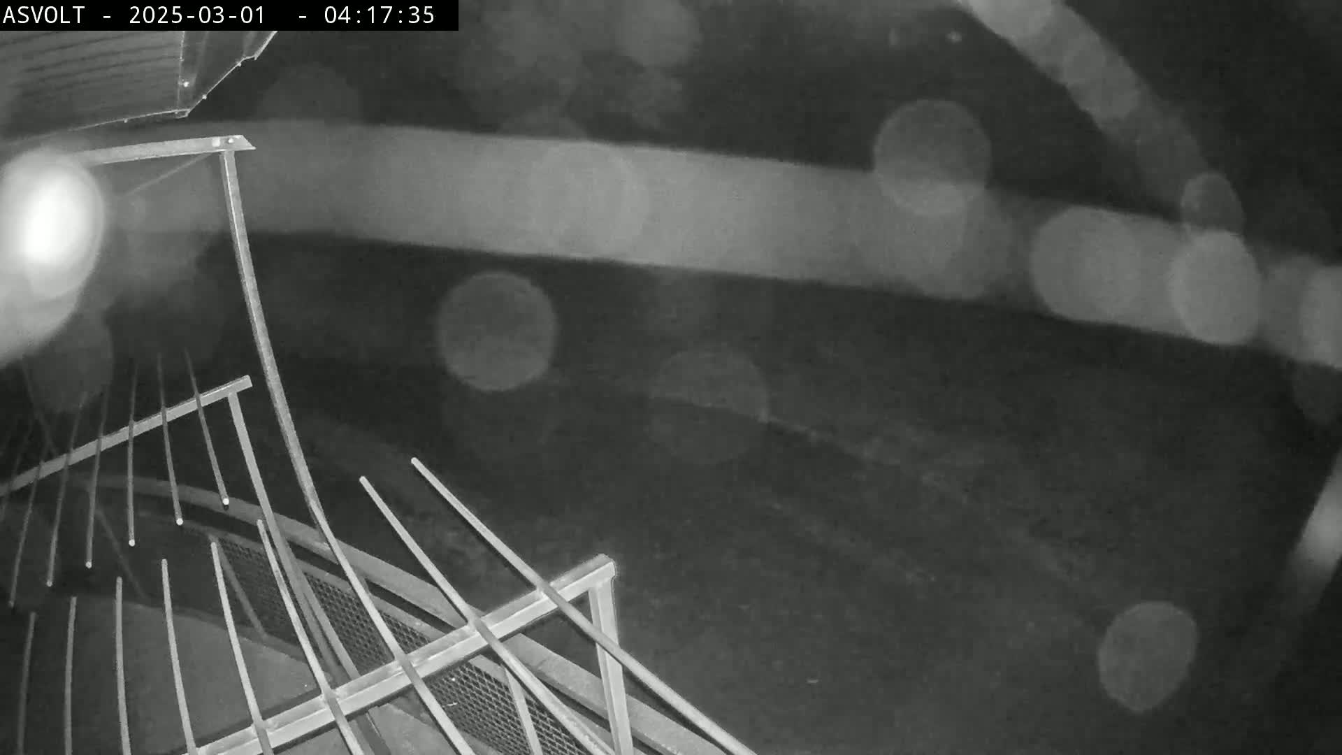A nighttime, rainy view shows a metal railing structure, possibly on a bridge or elevated platform.