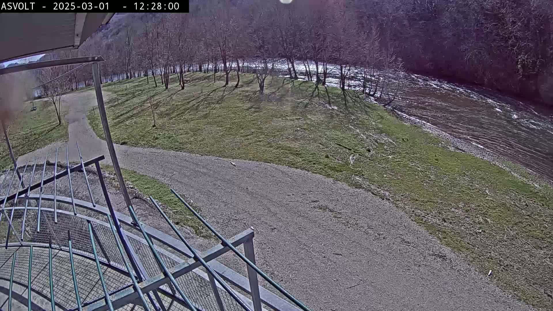 A sunny day overlooking a gravel path leading to a grassy area beside a flowing river bordered by bare trees.