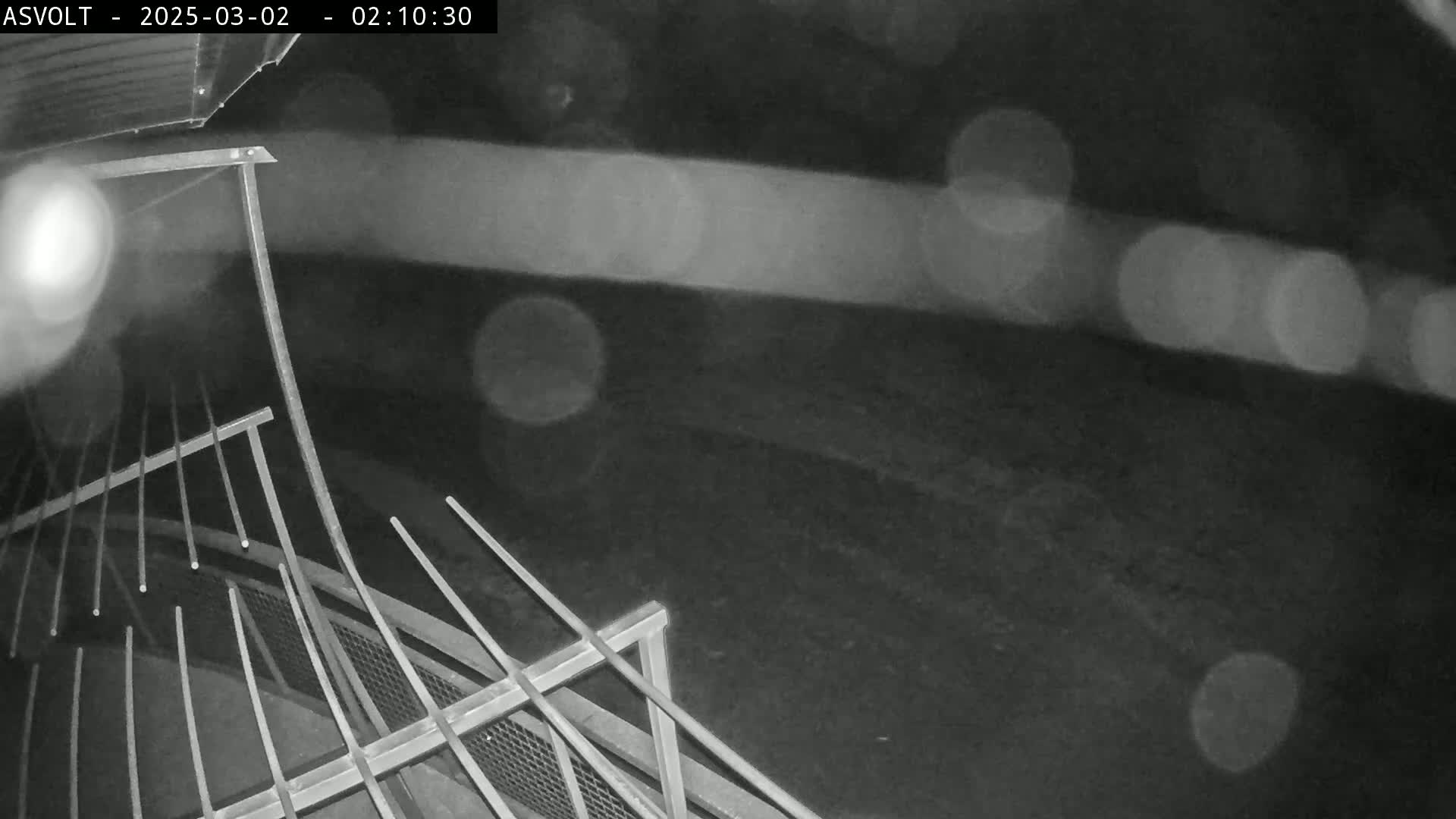 A nighttime, rainy view of a metal railing and a dark, blurry ground.