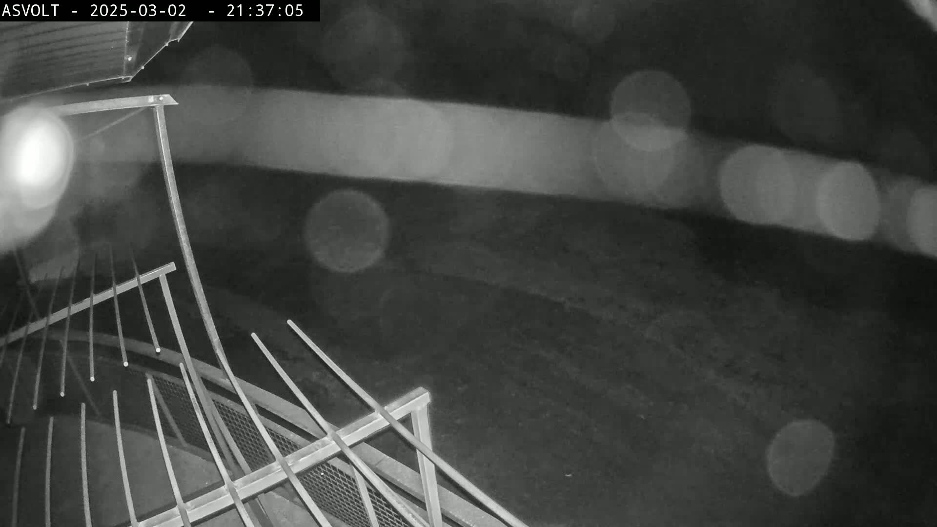 A nighttime, rain-blurred view shows a section of metal railing and fencing in the foreground with a dark, indistinct background.