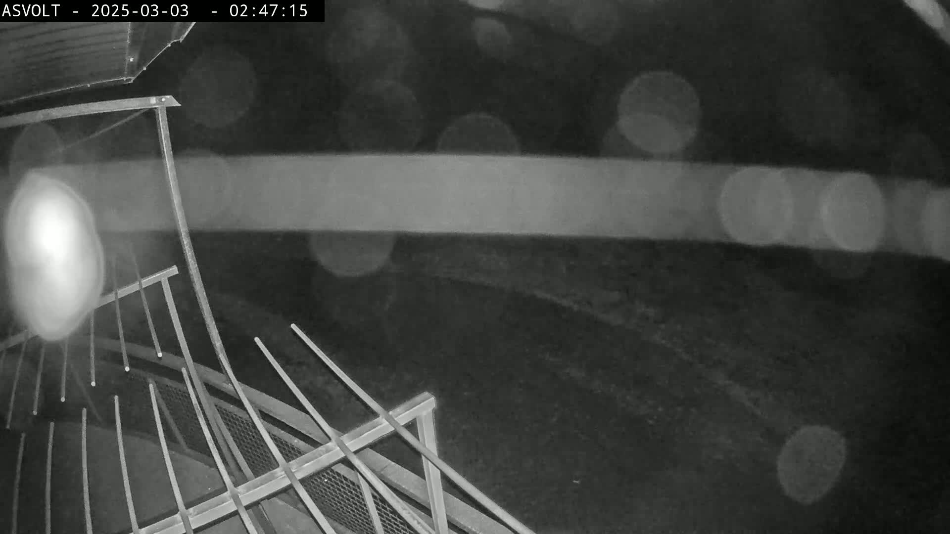 A nighttime, blurry, black-and-white image shows a metal railing overlooking a dark area with blurred lights in the background, possibly due to rain or fog.