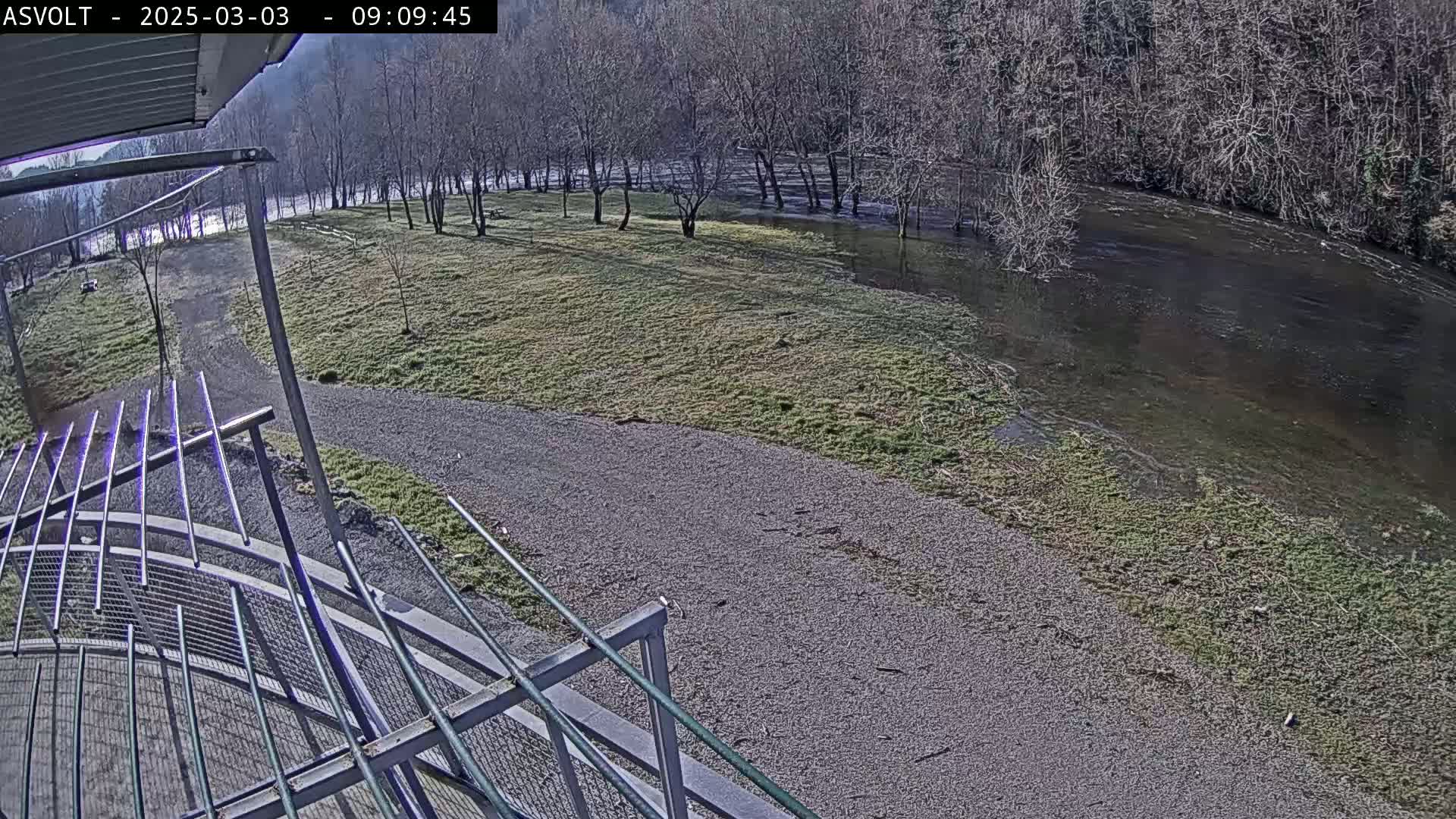 A gravel path leads to a riverbank where the water has overflowed onto a grassy area, under a sunny sky.