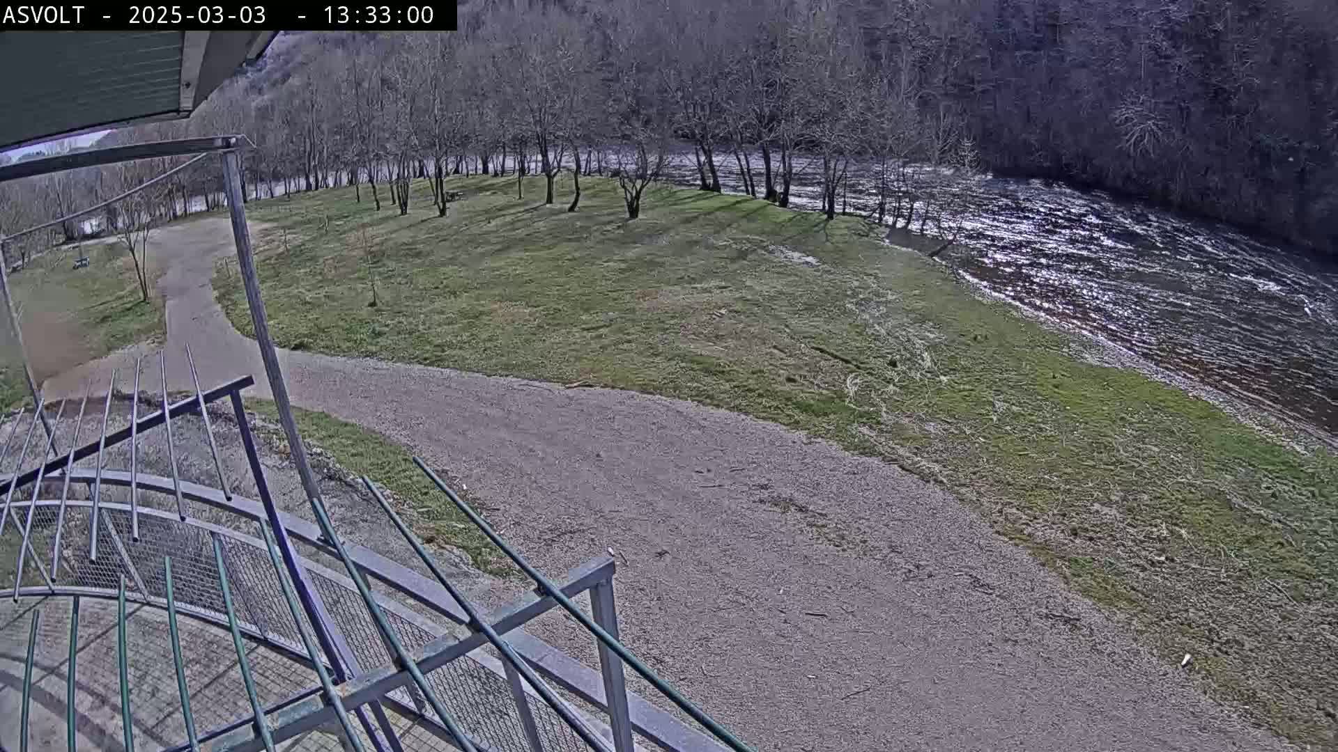 A gravel path leads to a grassy area beside a flowing river, under a mostly sunny sky, with bare trees lining the riverbank.