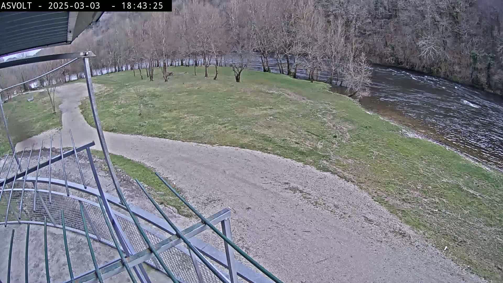 A gravel path curves near a grassy area next to a river, with bare trees lining the riverbank under a cloudy sky.