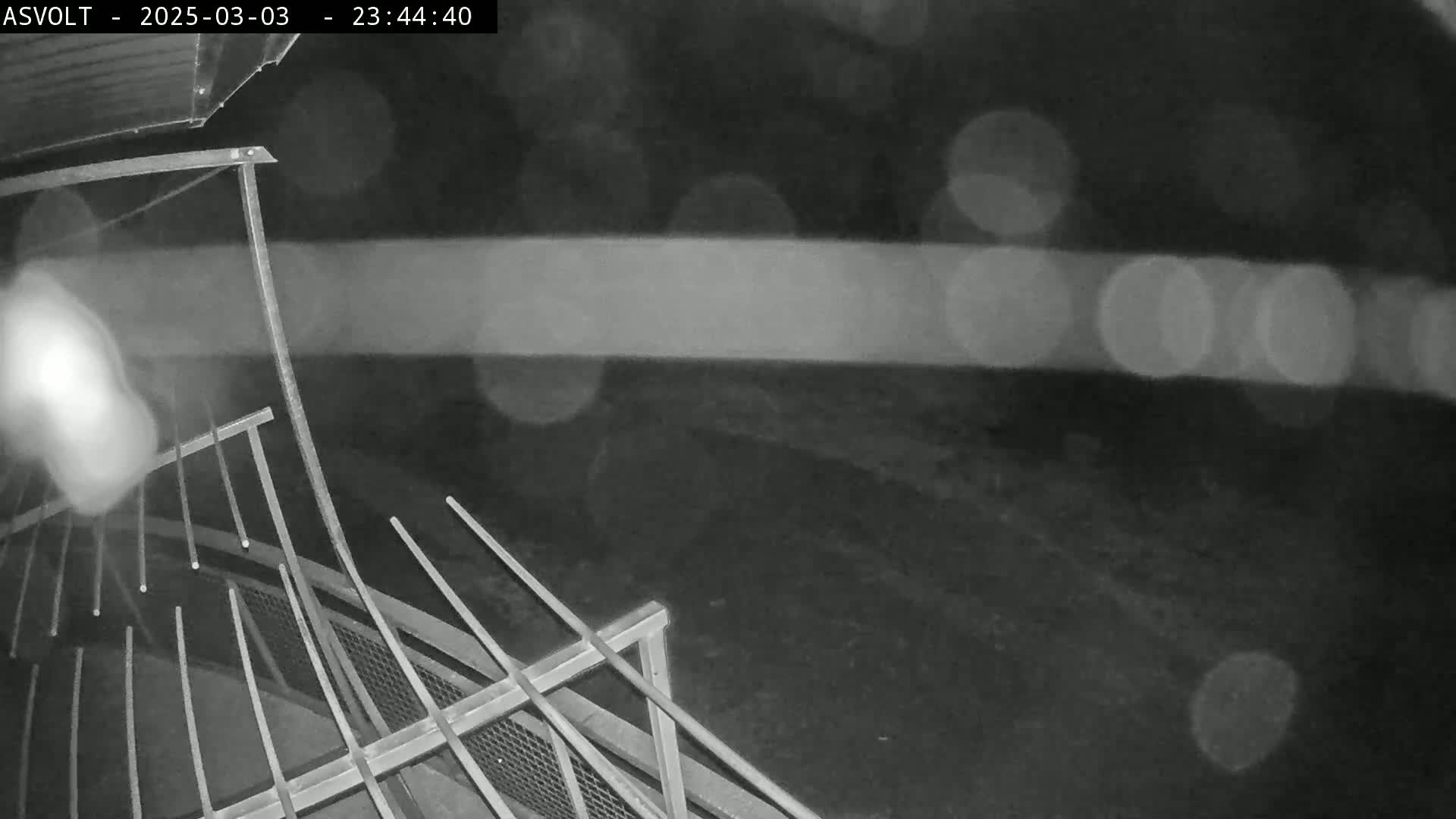 A nighttime, rainy scene shows a portion of a metal railing and a dark, blurry ground.