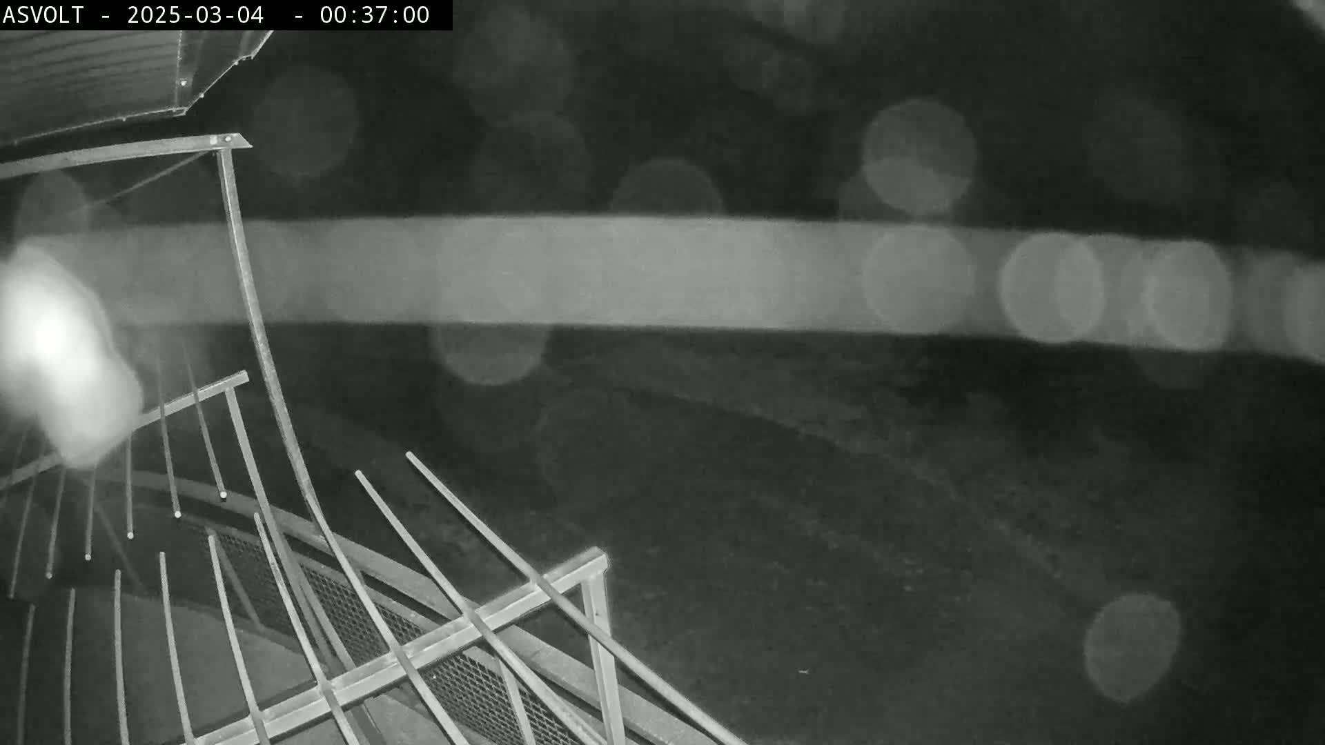 A nighttime, blurry view from above shows a metal railing overlooking a dark area with blurred lights in the background.