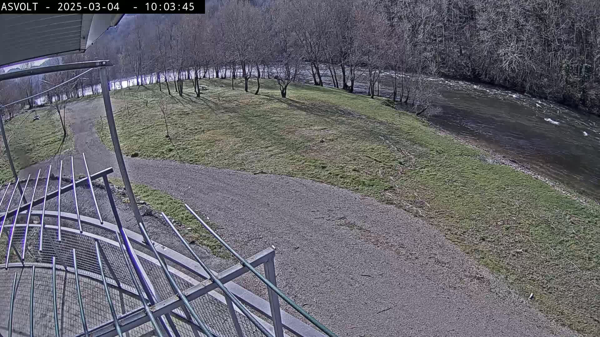 A gravel path leads to a grassy area beside a flowing river under a mostly sunny sky, viewed from an elevated position with a metal railing in the foreground.