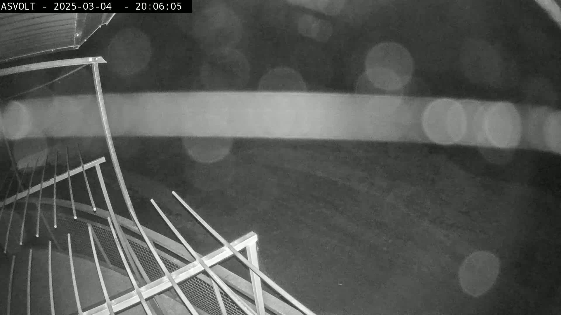 A nighttime, blurry black-and-white image shows a metal structure, possibly a railing or fence, with a dark, indistinct background and blurry lights in the distance.