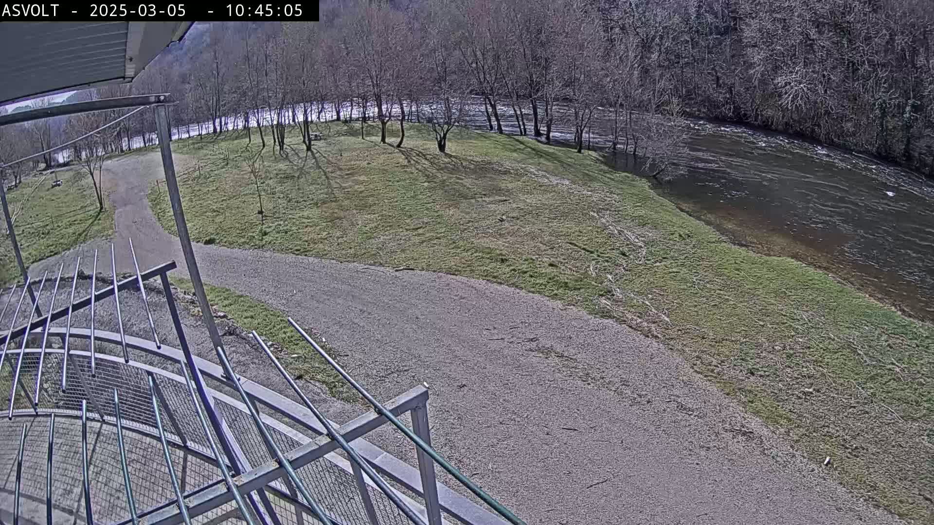 A gravel path curves toward a riverbank under a partly sunny sky, with bare trees lining the river and sparse grass covering the ground.