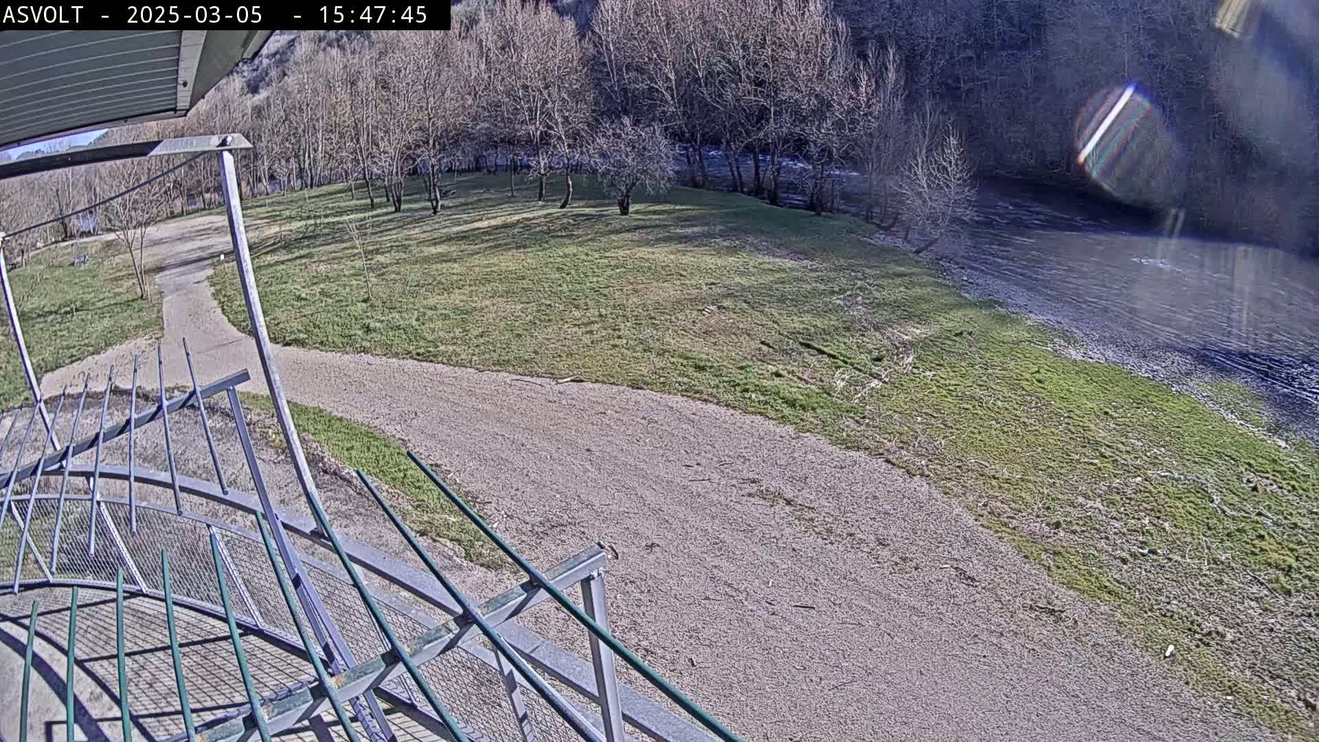 A sunny view from an elevated position shows a gravel path leading to a grassy area beside a river, partially obscured by a metal railing in the foreground.