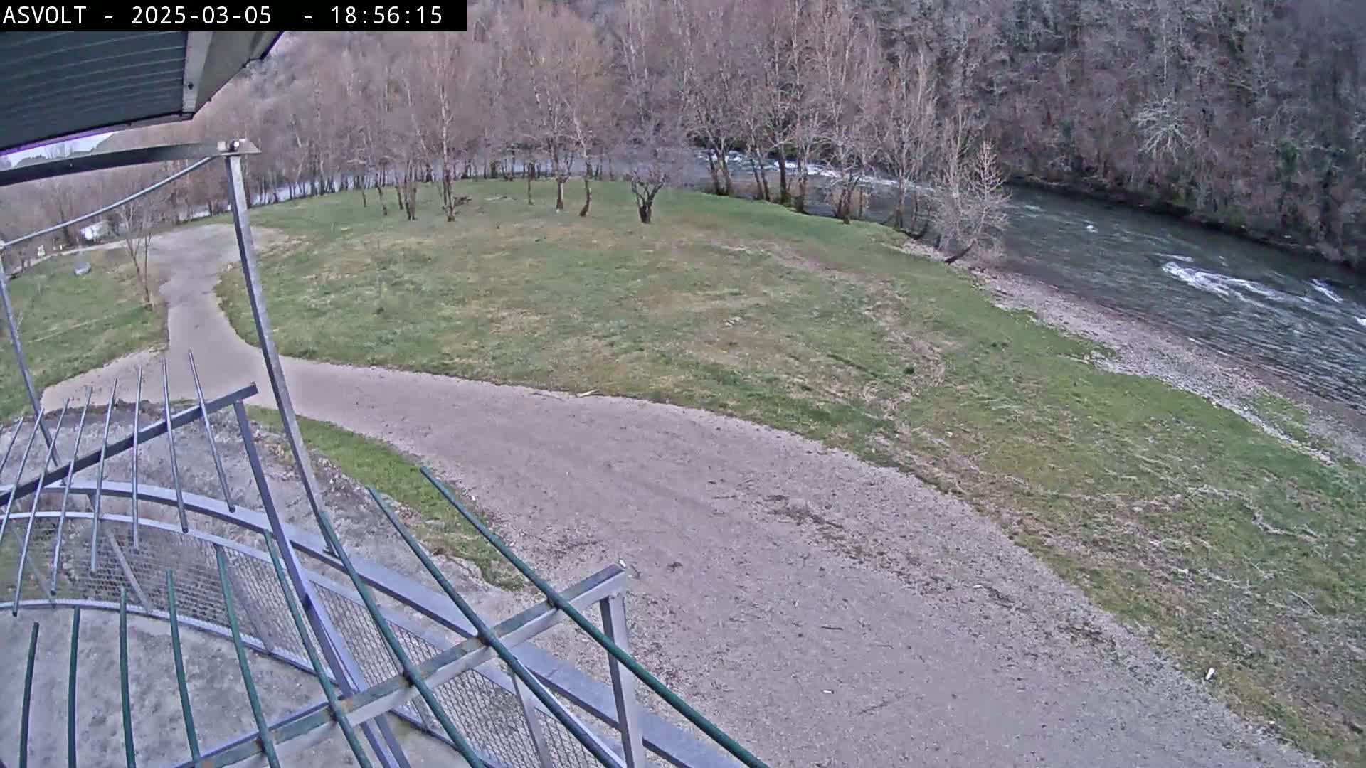 A view from an elevated position shows a gravel path leading to a grassy area next to a river under a cloudy sky, with bare trees lining the riverbank.