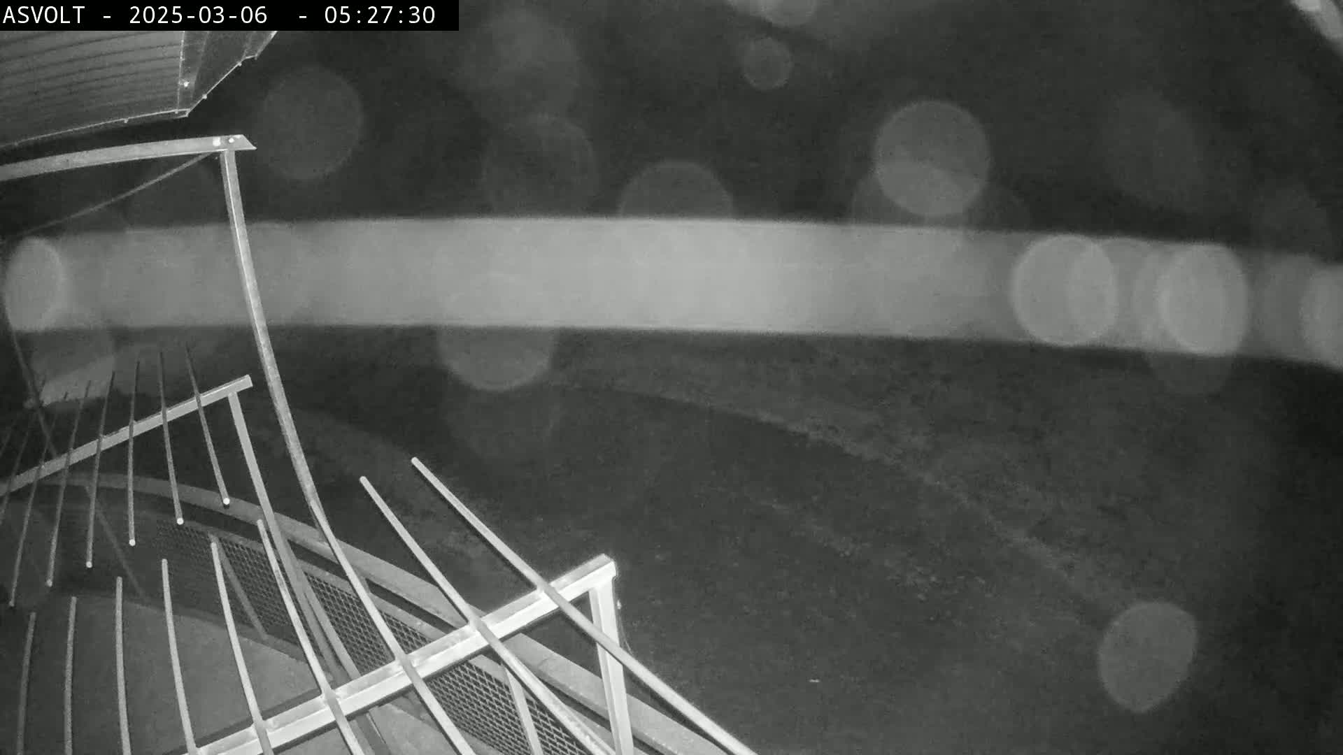 A nighttime, blurry image shows a metal structure resembling a railing or fence, with blurred lights visible in the background under light rain.