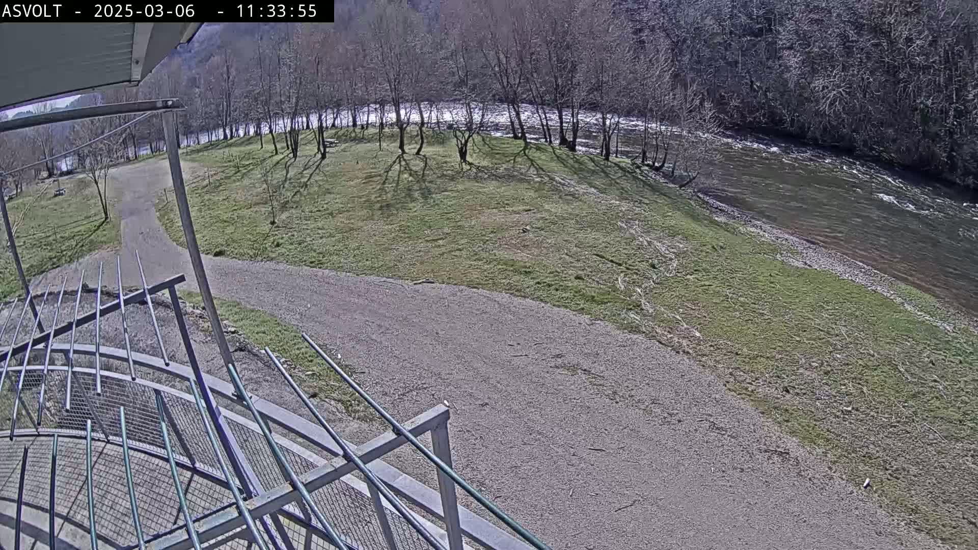 A gravel path leads to a grassy area beside a river under a sunny sky, viewed from an elevated position behind a metal railing.