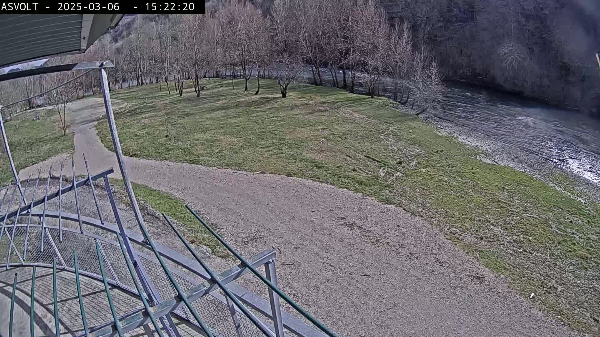 A gravel path winds through a grassy area toward a river under a mostly sunny sky, next to a metal railing.