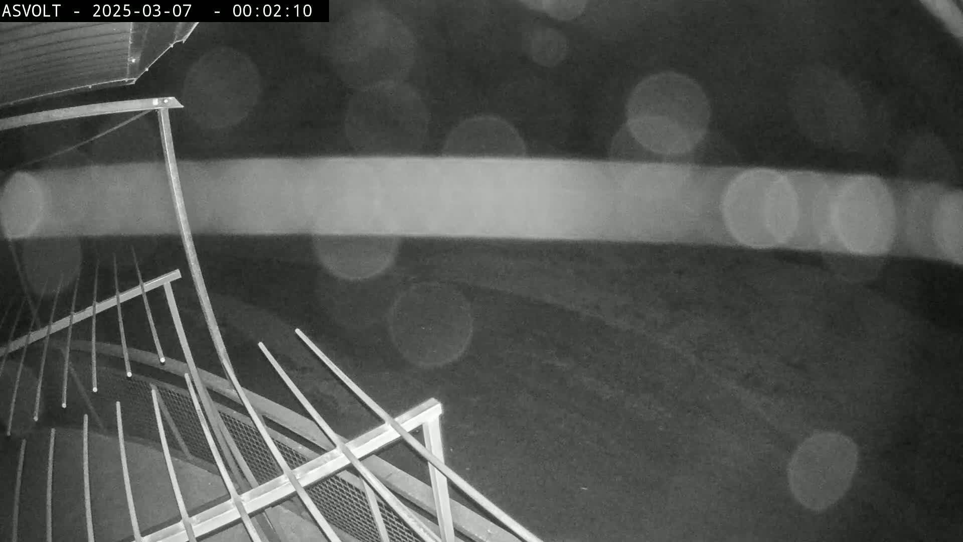 A nighttime, blurry outdoor view shows a metal railing and a section of ground, with water droplets visible on the lens.