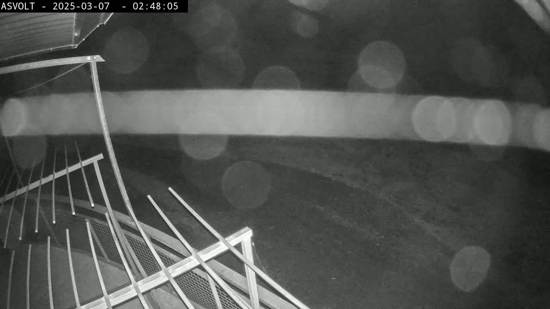 A nighttime, blurry, black-and-white image shows a metal railing, possibly part of a staircase or fence, in the rain.