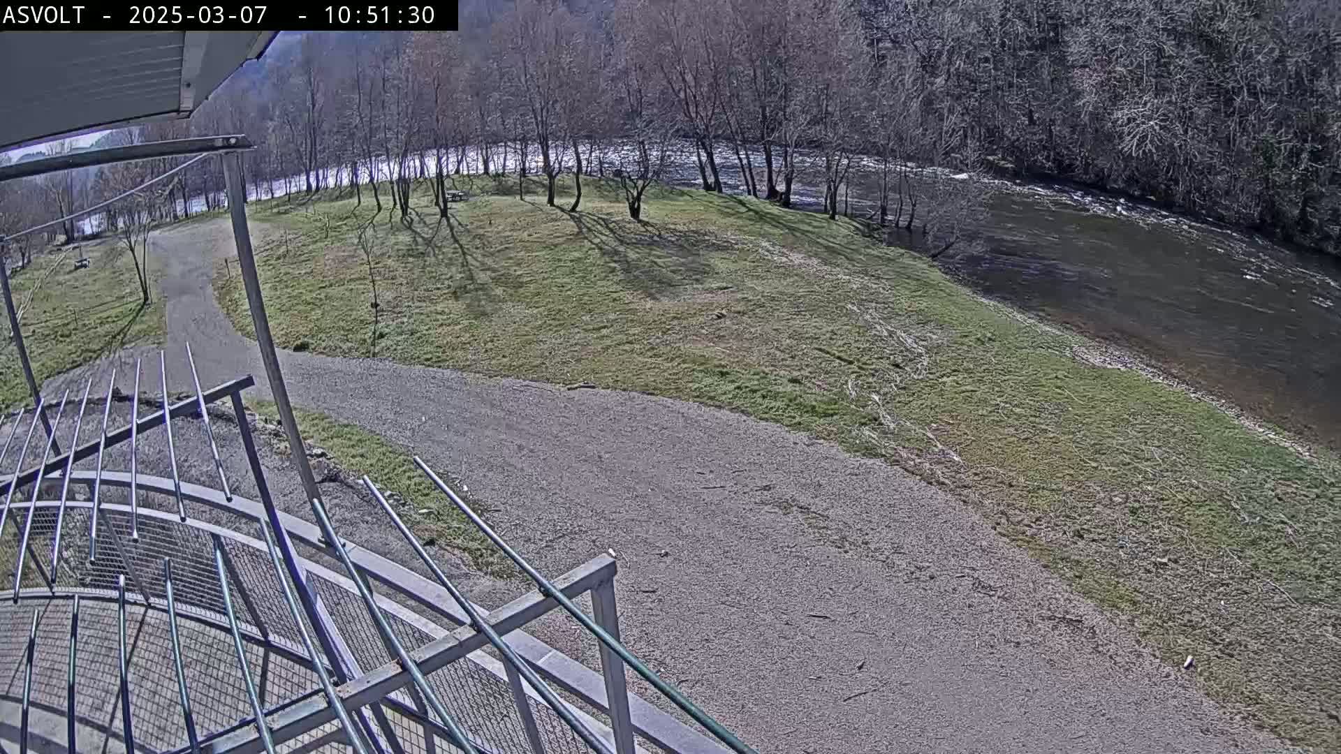 A gravel path leads to a grassy area beside a river under a sunny sky, with bare trees lining the riverbank in the background.