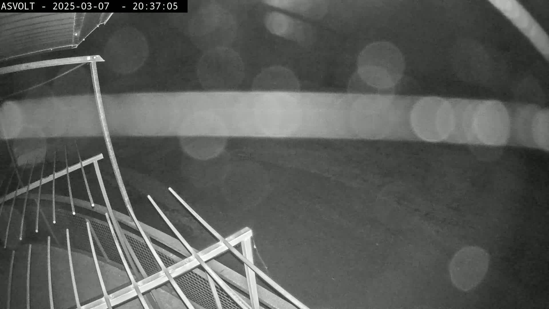 A nighttime, rainy view of a metal railing and a dark ground.
