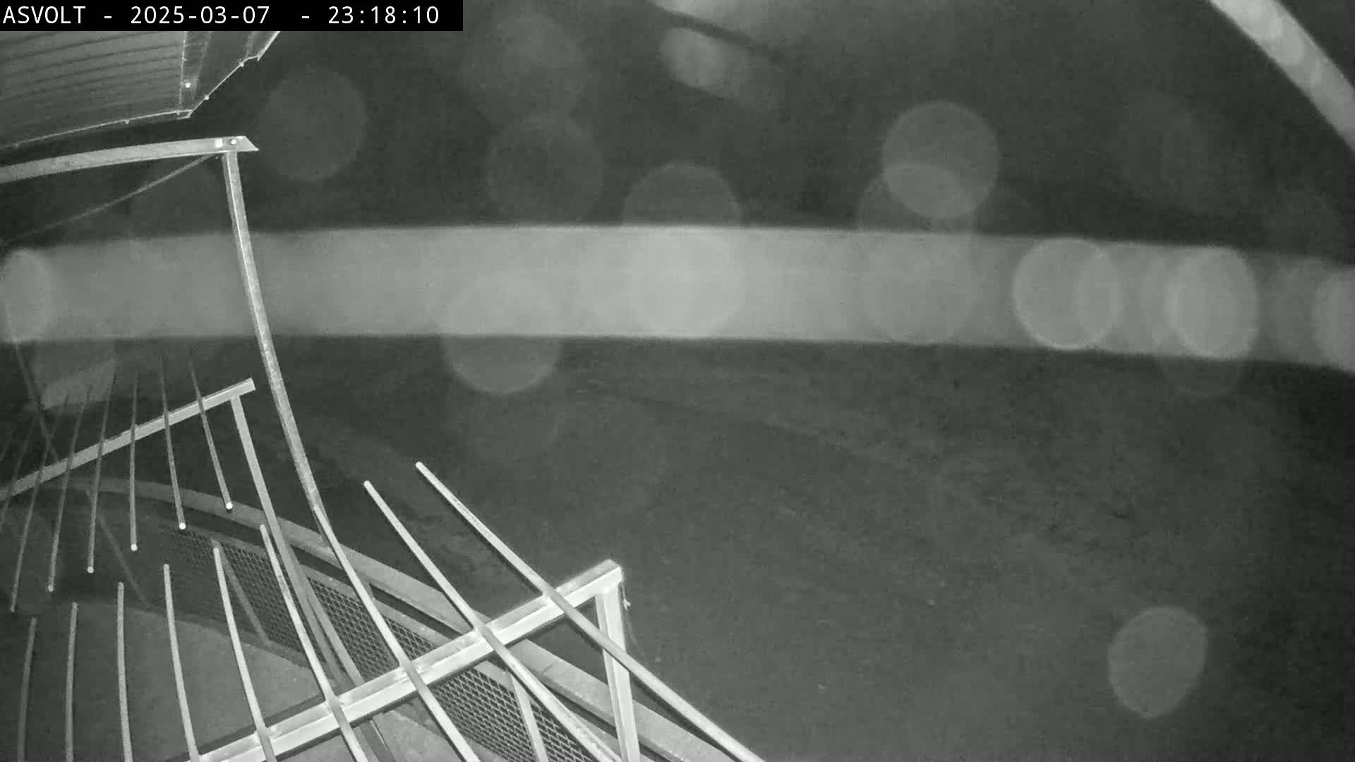 A nighttime, rainy scene shows a metal railing and a portion of a dark, flat surface outside.