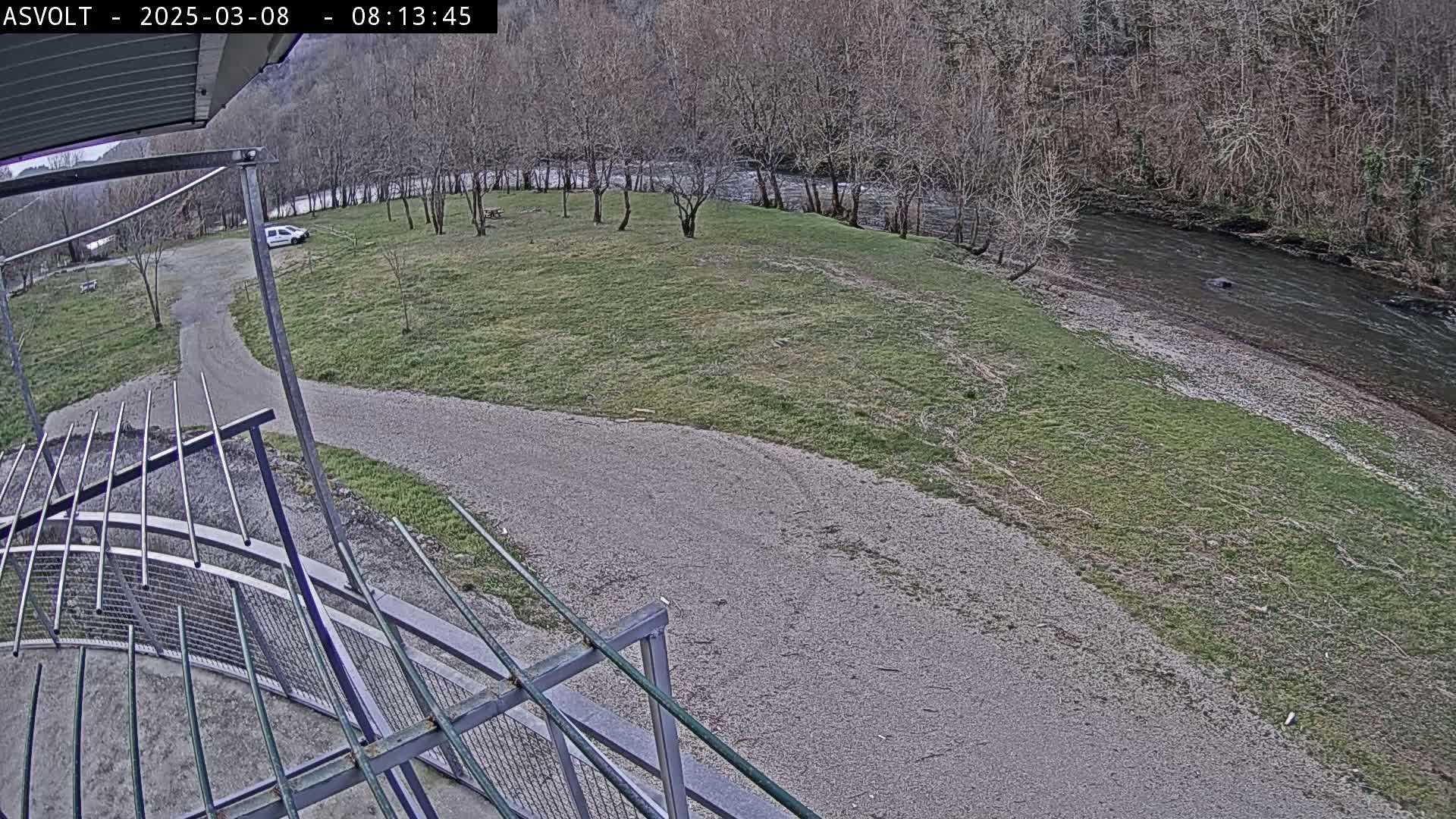 A view from an elevated vantage point shows a gravel path curving toward a riverbank, with a grassy area and a few bare trees in the background under an overcast sky.