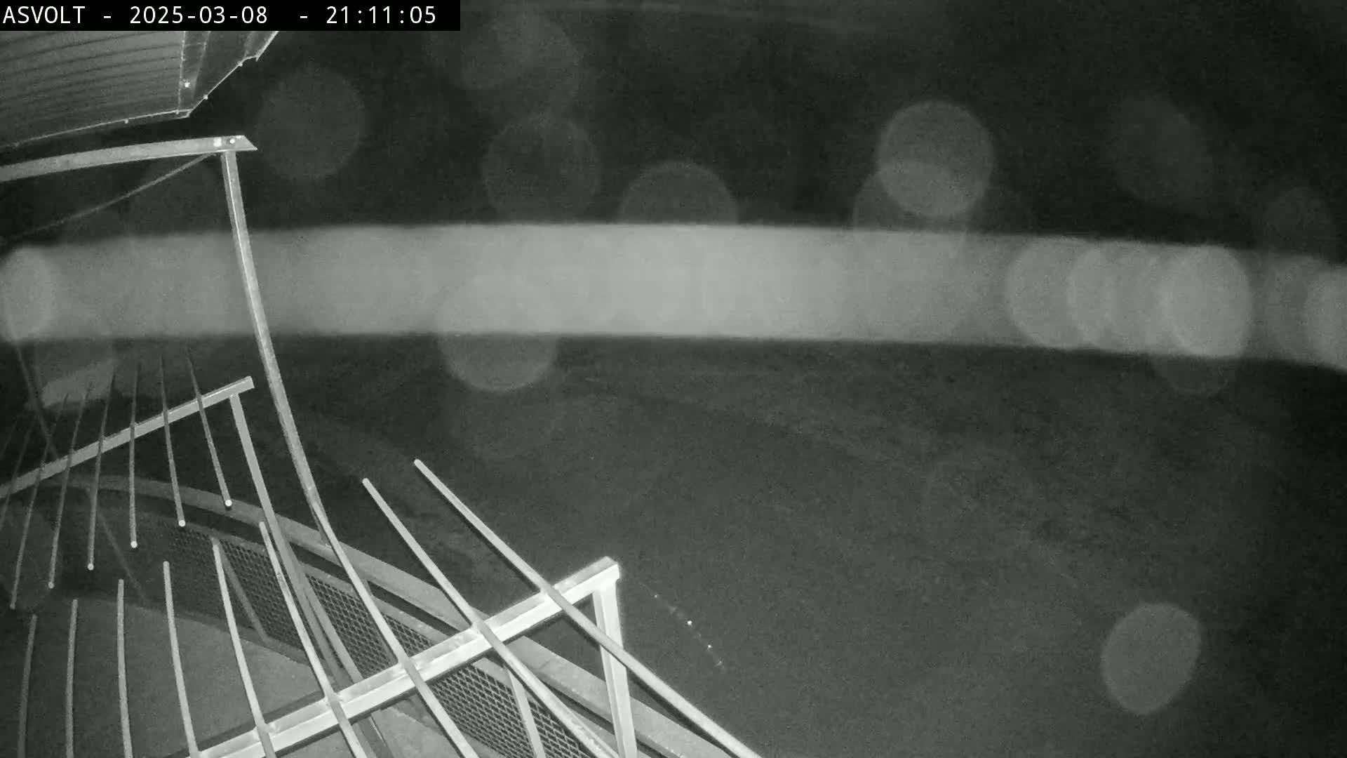 A nighttime, blurry, black-and-white image shows a metal railing overlooking a dark ground with blurred lights in the background.