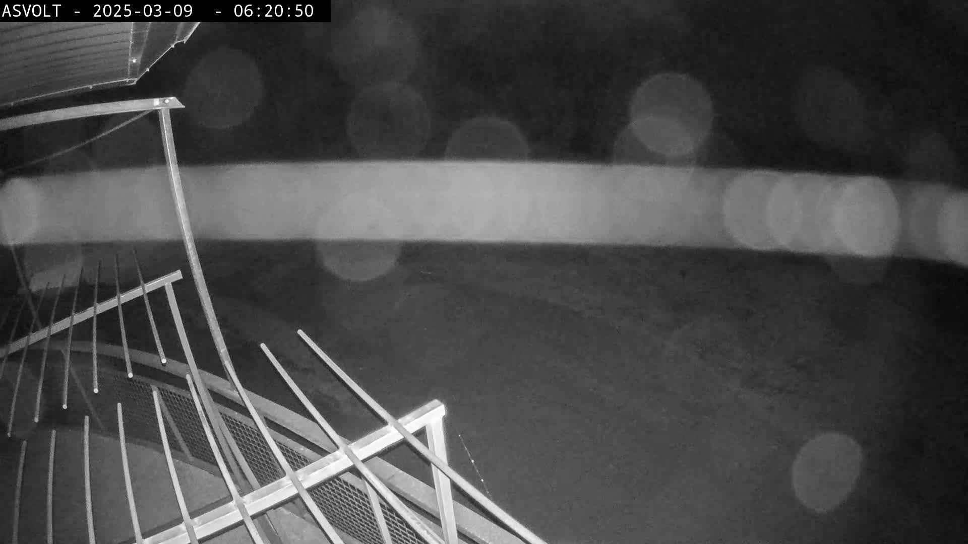 A nighttime, rainy scene showing a portion of a metal railing and a dark ground below, possibly from a security camera.