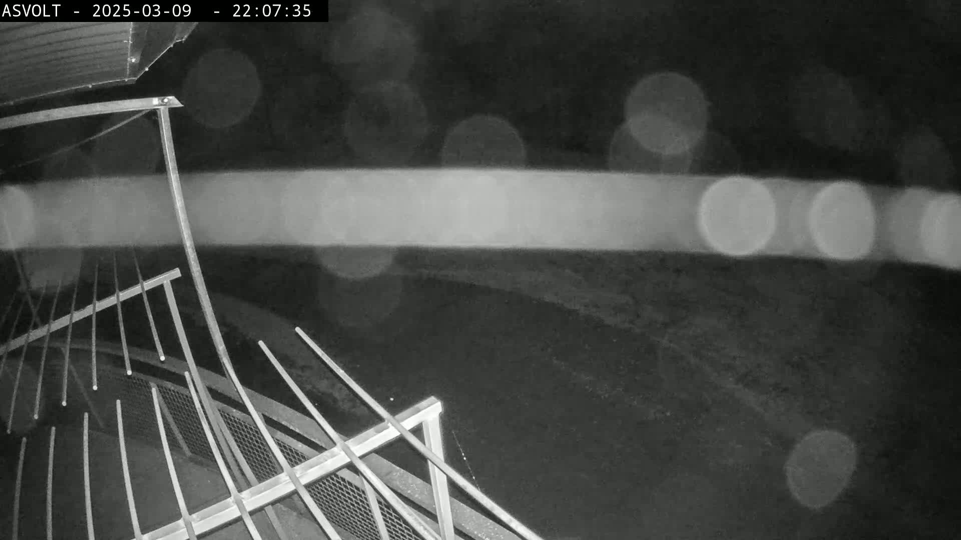 A nighttime, low-angle view shows a metal railing and fence in the rain with blurred lights in the distance.