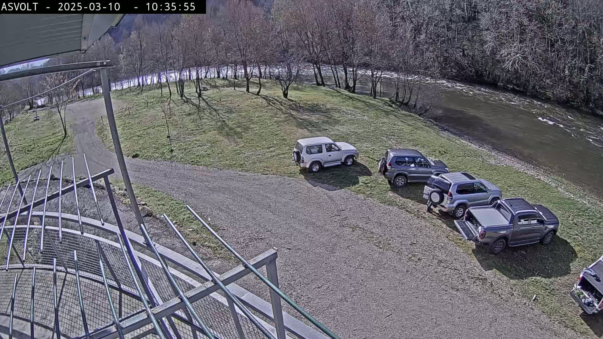 Four vehicles are parked in a gravel lot next to a grassy area and a river, under a mostly sunny sky.