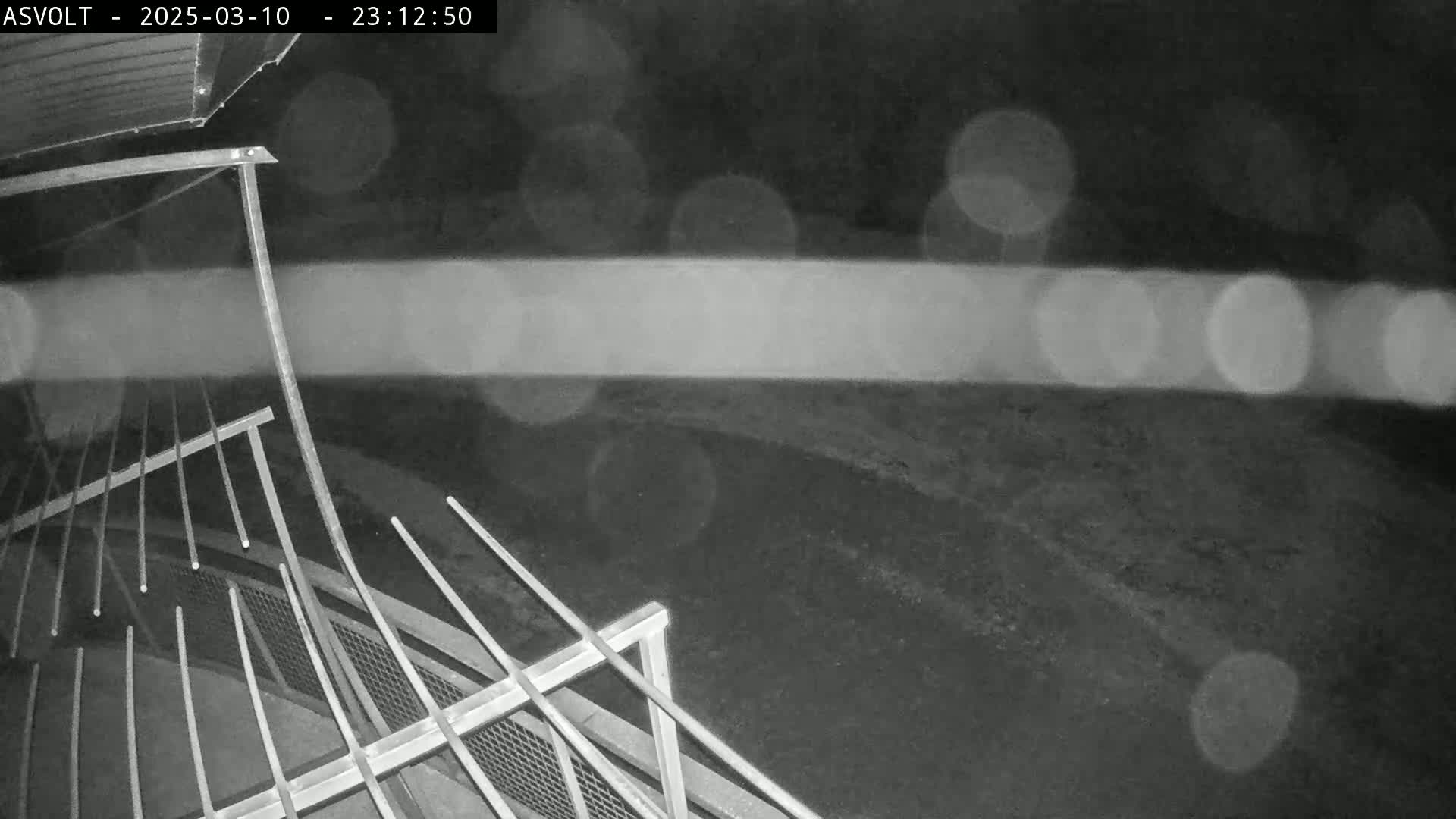 A nighttime, rainy view from an elevated angle shows a metal railing and a dark, blurry paved area beyond.