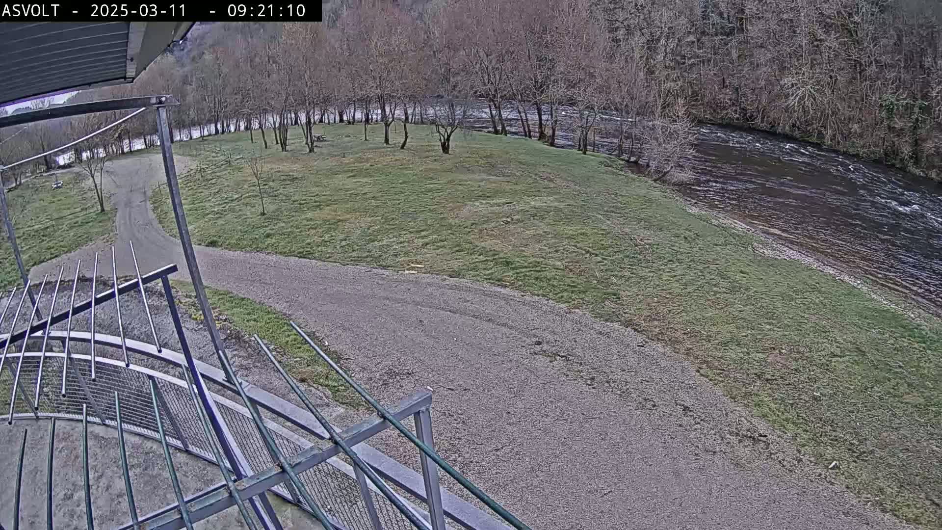 A gravel path leads to a grassy area next to a river under an overcast sky, viewed from an elevated perspective.