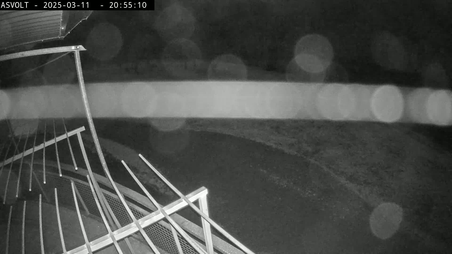 A nighttime, blurry image shows a section of metal railing overlooking a dark area with blurred lights in the background, possibly due to rain or fog.