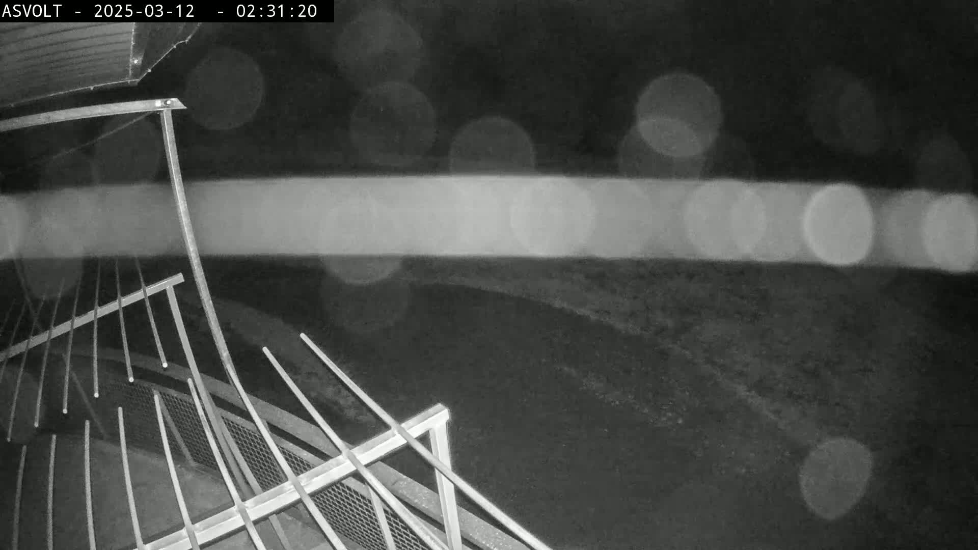 A nighttime, rain-blurred view shows a metal railing structure overlooking a dark, paved area.
