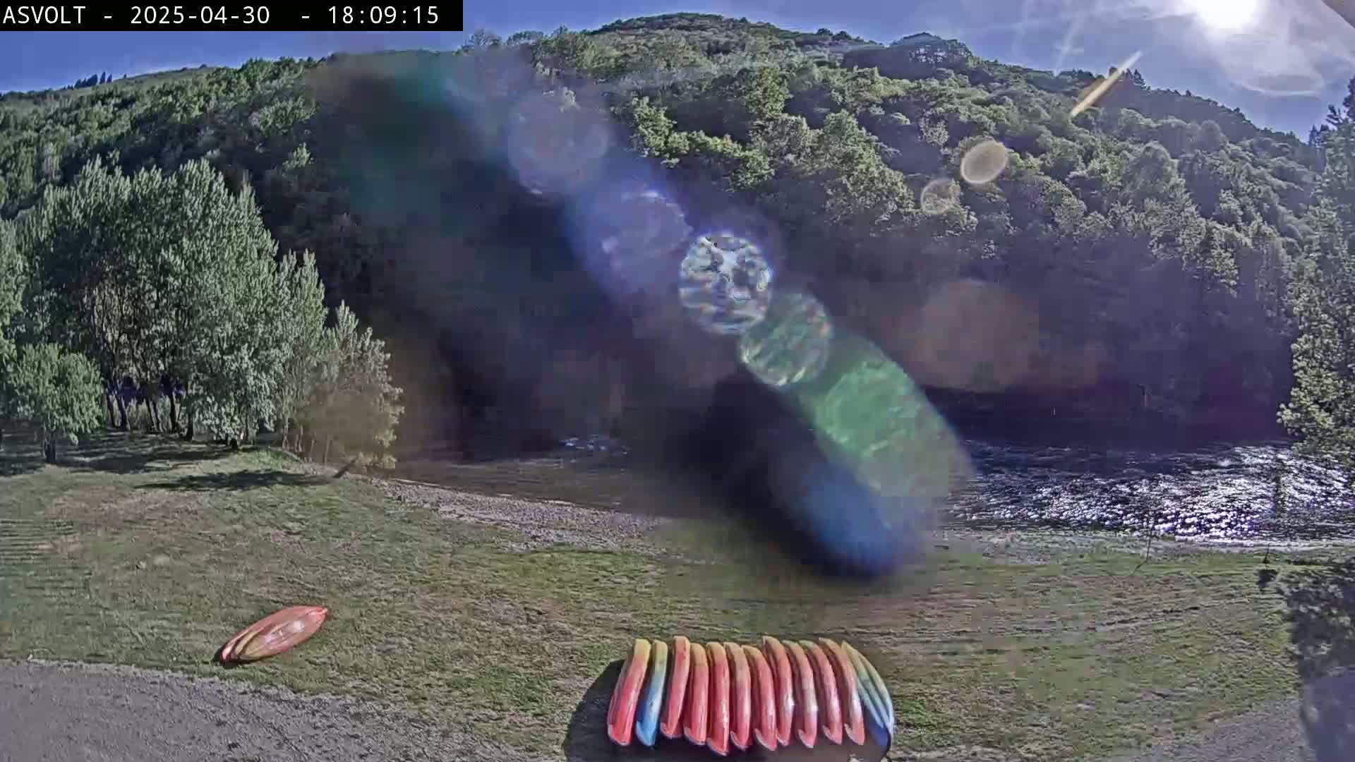 Kayaks are on the grassy bank of a river under a sunny sky, with lush green hills in the background.