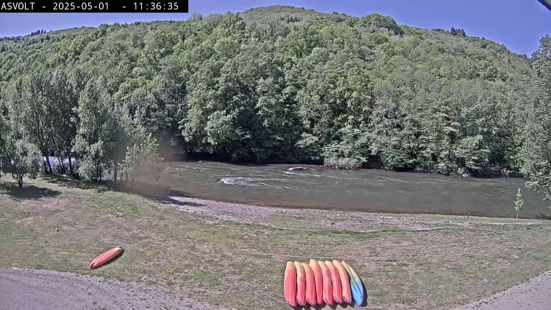 A river flows under a sunny sky past a grassy bank where several kayaks are resting.