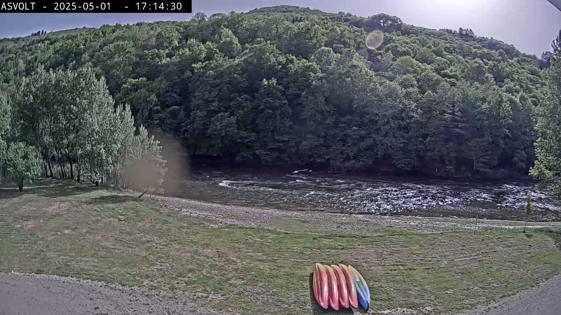 Several kayaks lay on a grassy bank overlooking a gently flowing river, surrounded by lush green trees under a partly sunny sky.