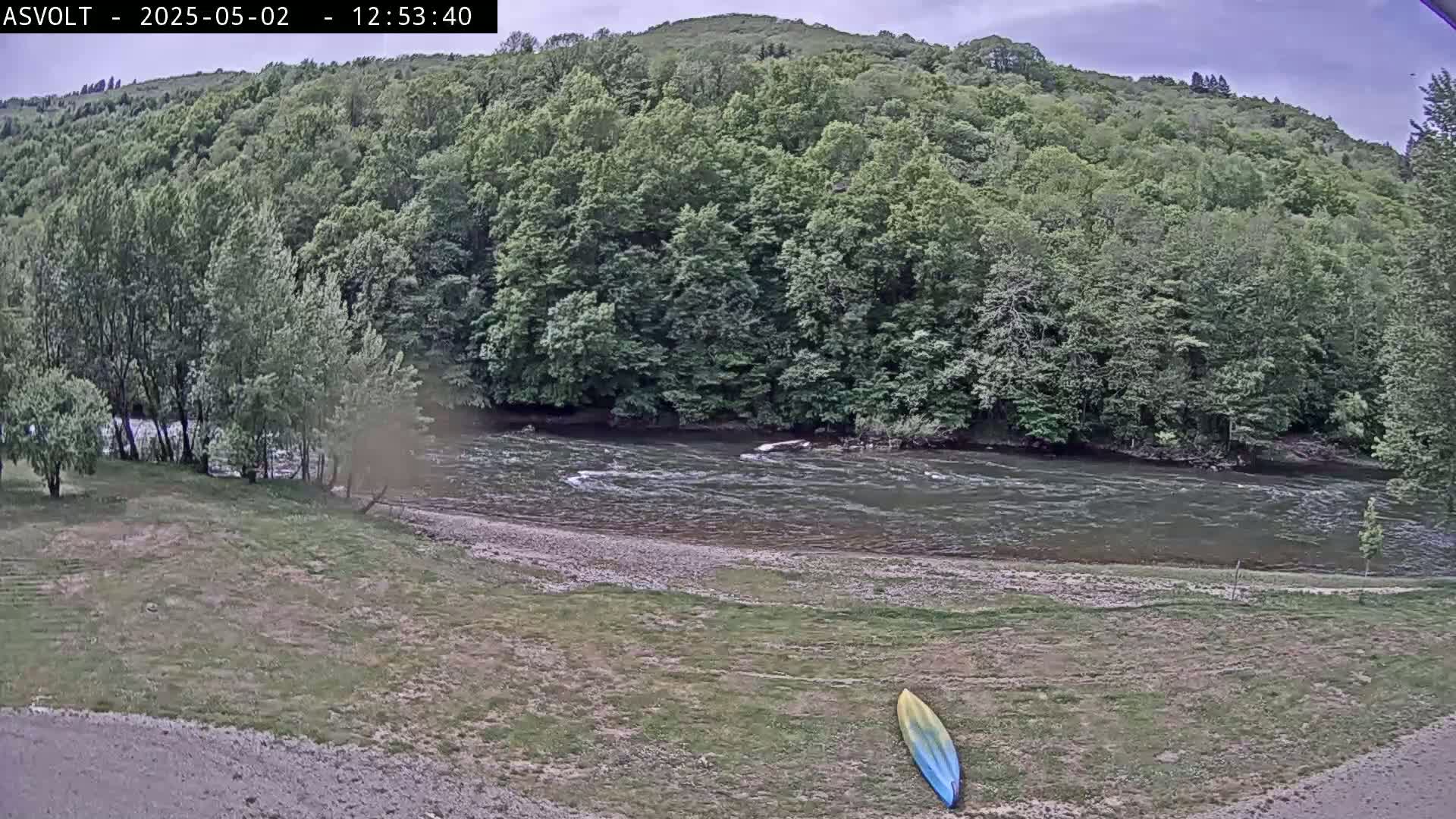 A kayak lies on the grassy bank of a river flowing through a lush green forest under a cloudy sky.