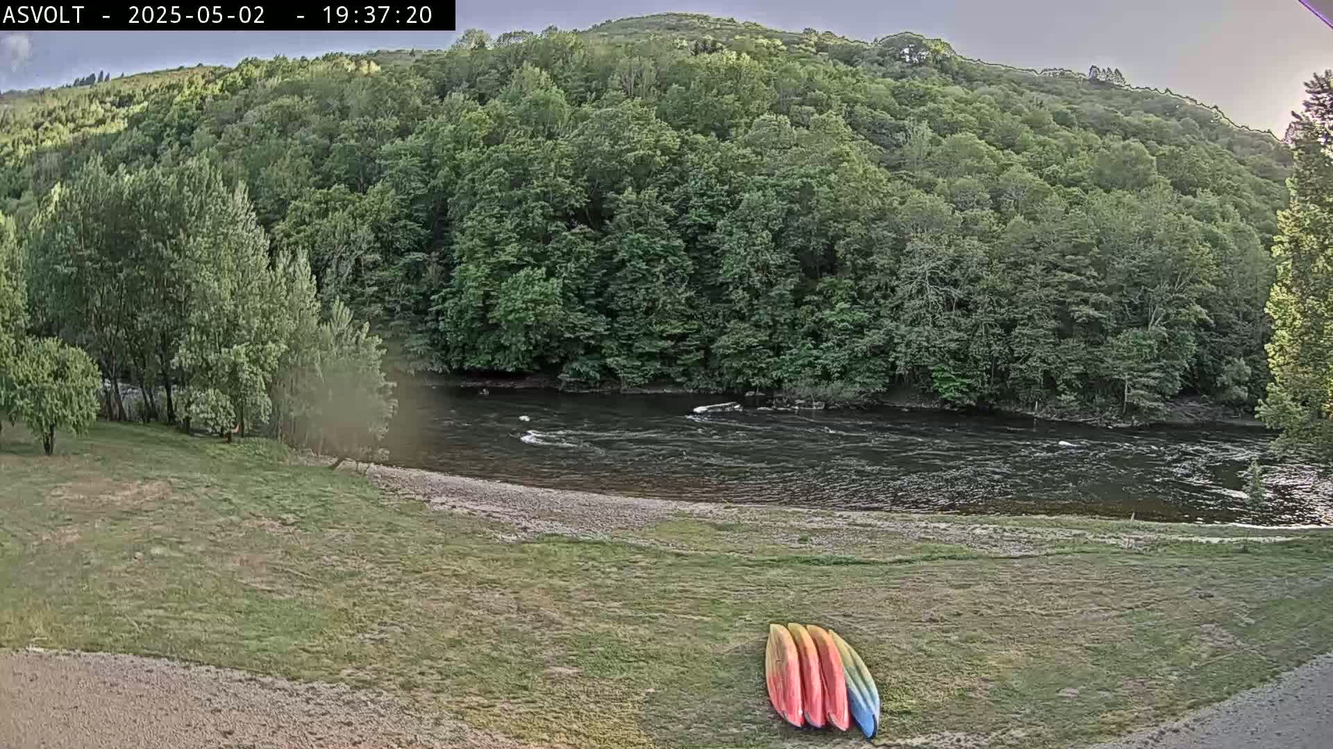 Several kayaks lay on a grassy bank overlooking a dark, gently flowing river bordered by lush green trees under a mostly cloudy sky.