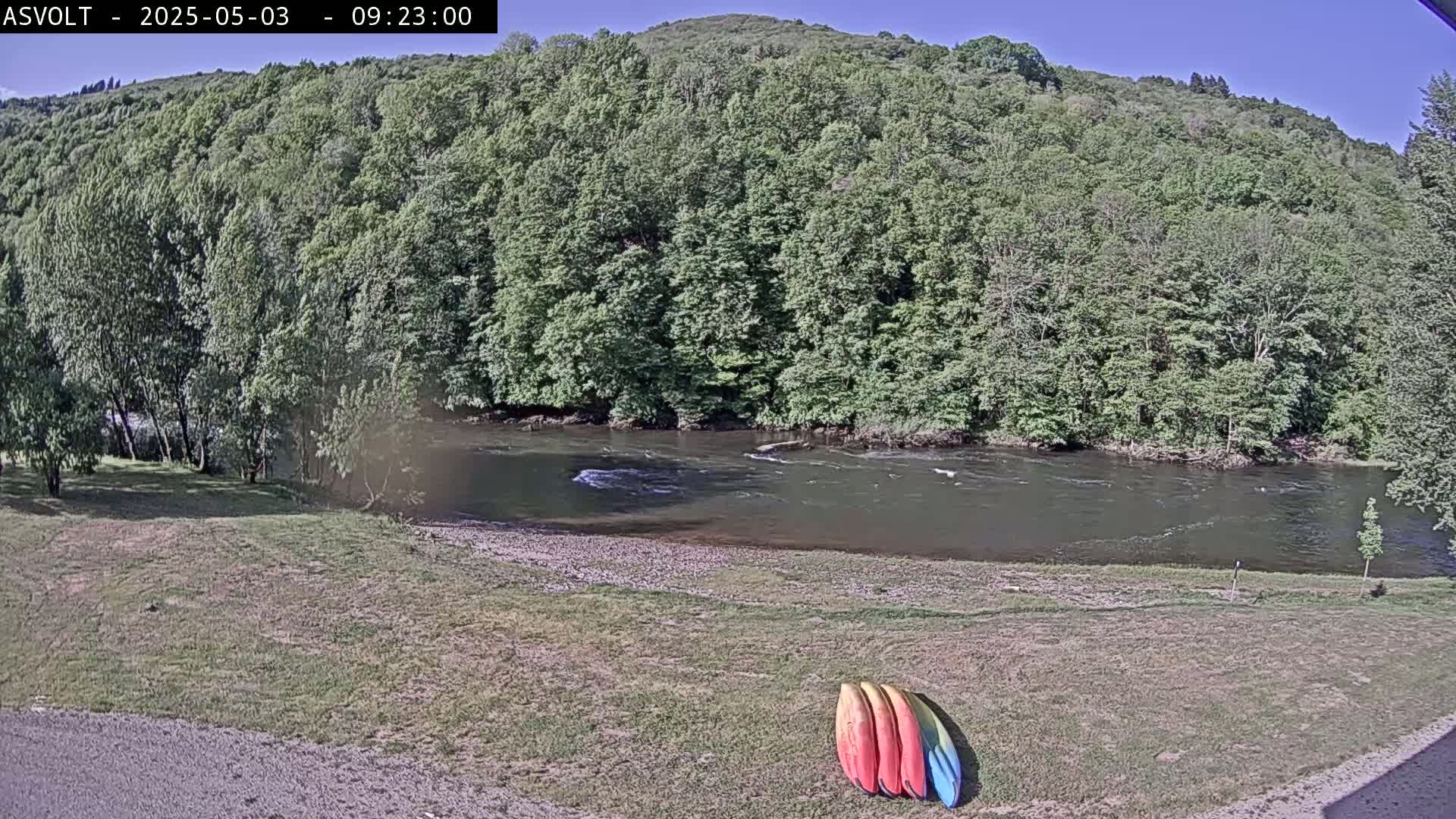 Three kayaks lie on a grassy area beside a calm river, which flows past lush green trees and a hillside under a clear blue sky.