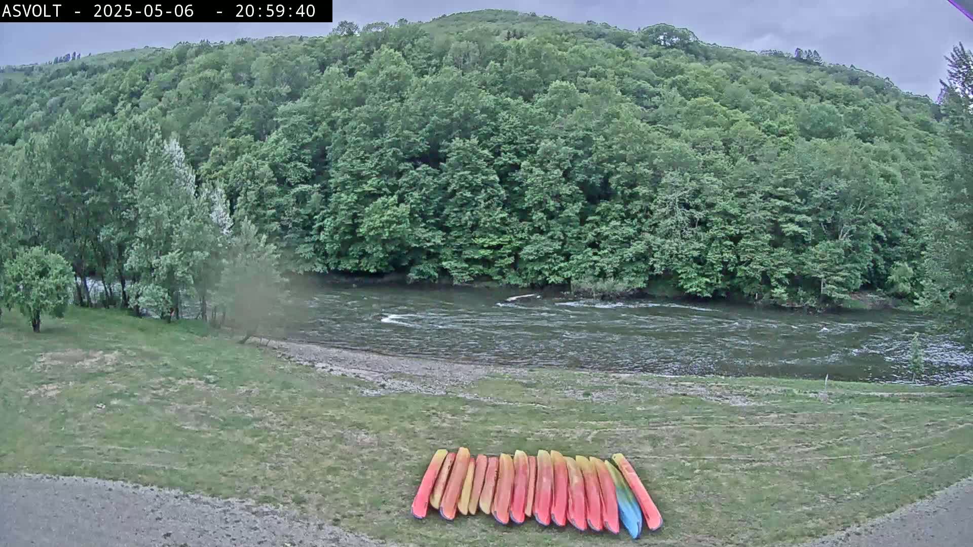 A river flows beneath a lush green hillside on an overcast day, with a row of colorful kayaks resting on the grassy riverbank.