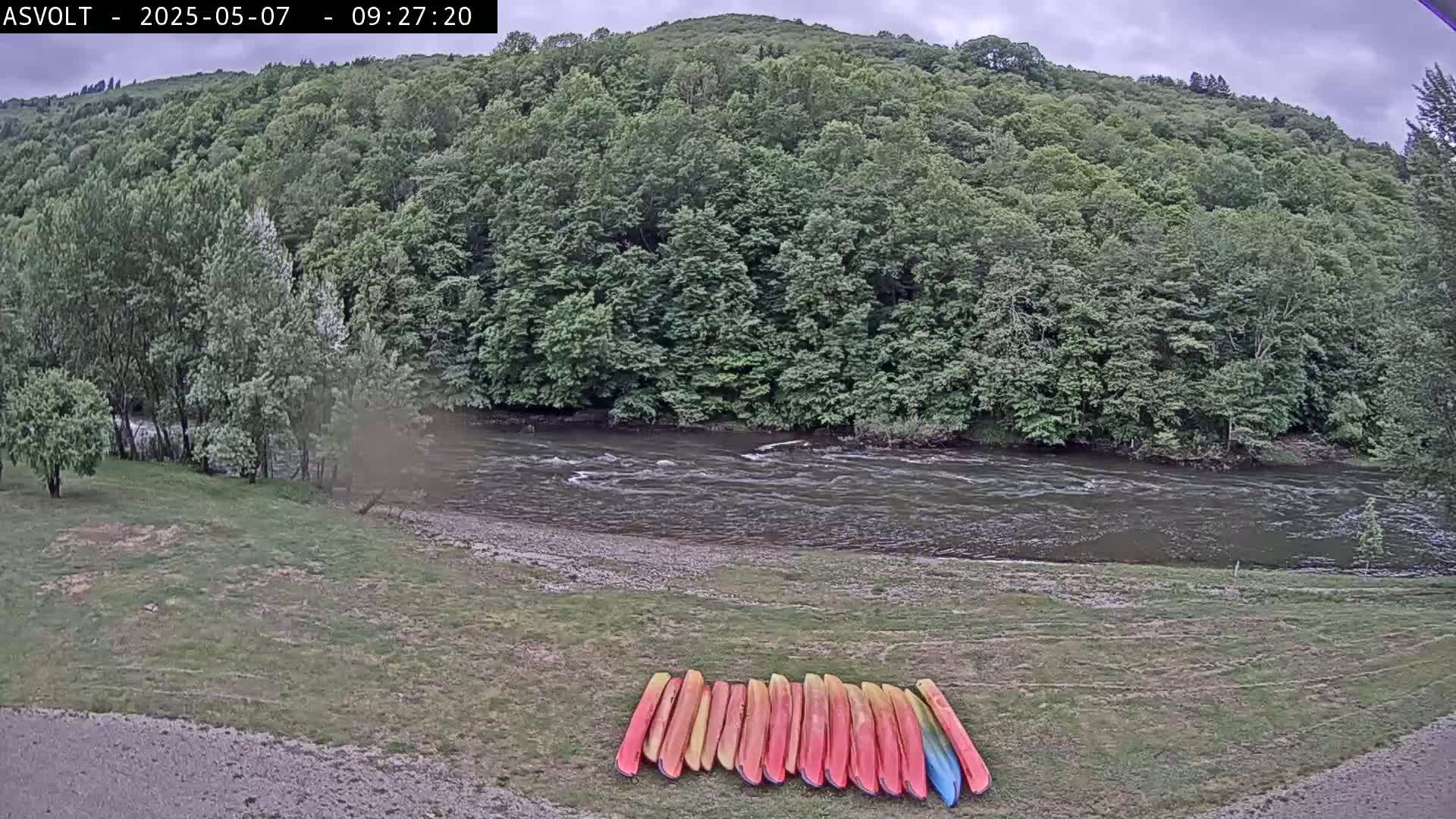 A row of kayaks rests on a grassy bank overlooking a flowing river, under an overcast sky, with lush green hills in the background.