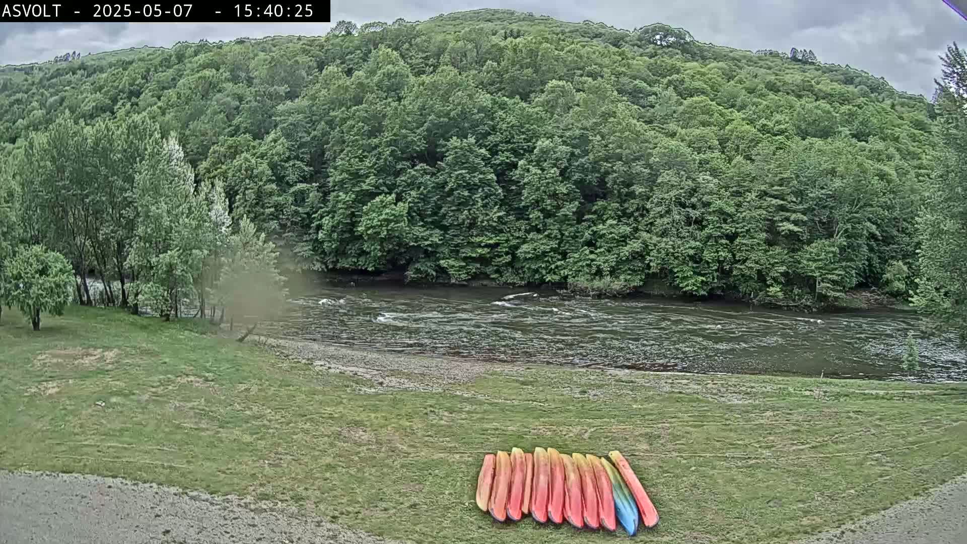 Several kayaks are lined up on a grassy bank beside a flowing river under a cloudy sky, with lush green trees forming a backdrop.