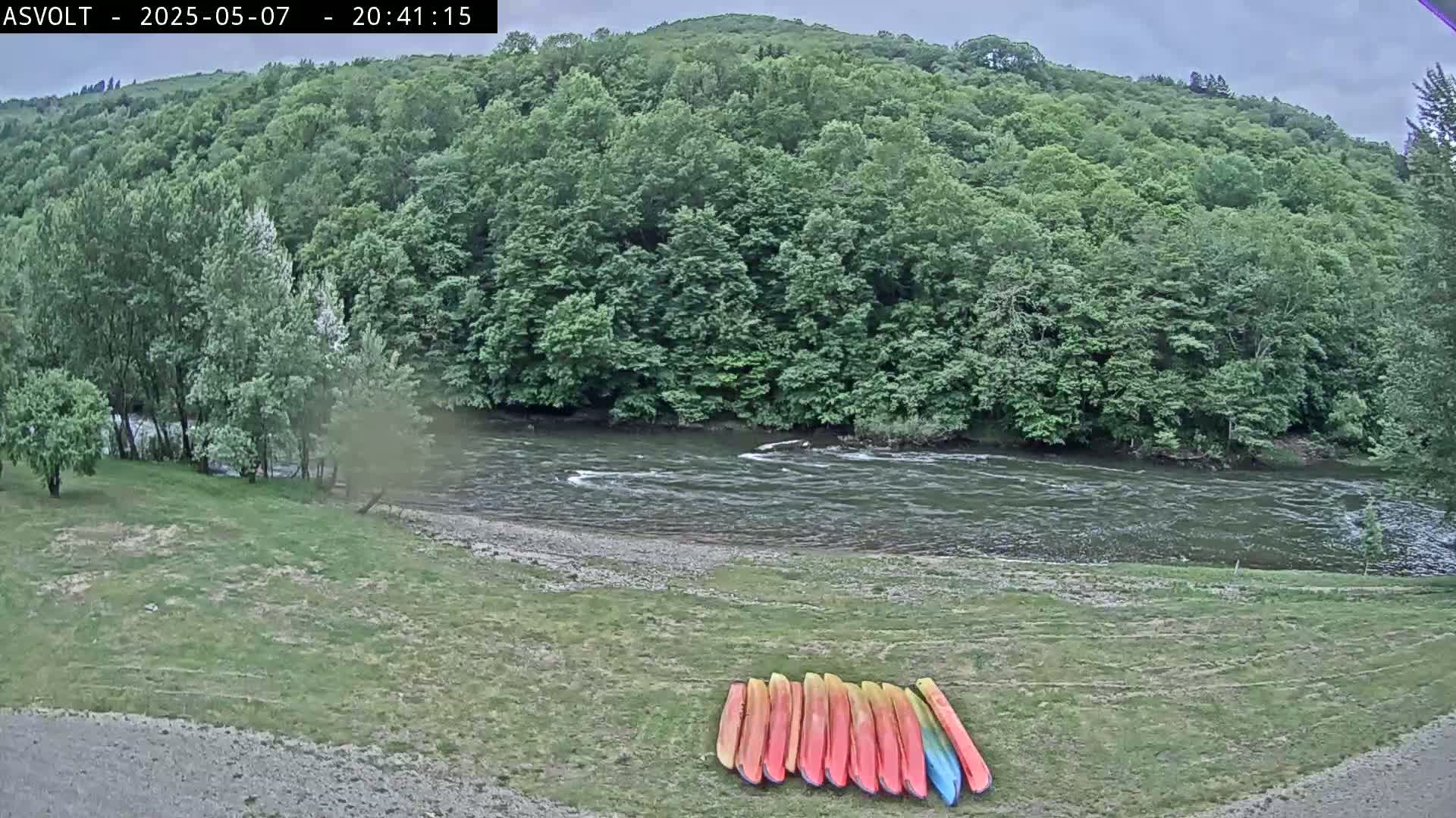 Several kayaks are lined up on a grassy riverbank, next to a flowing river surrounded by lush green trees under an overcast sky.