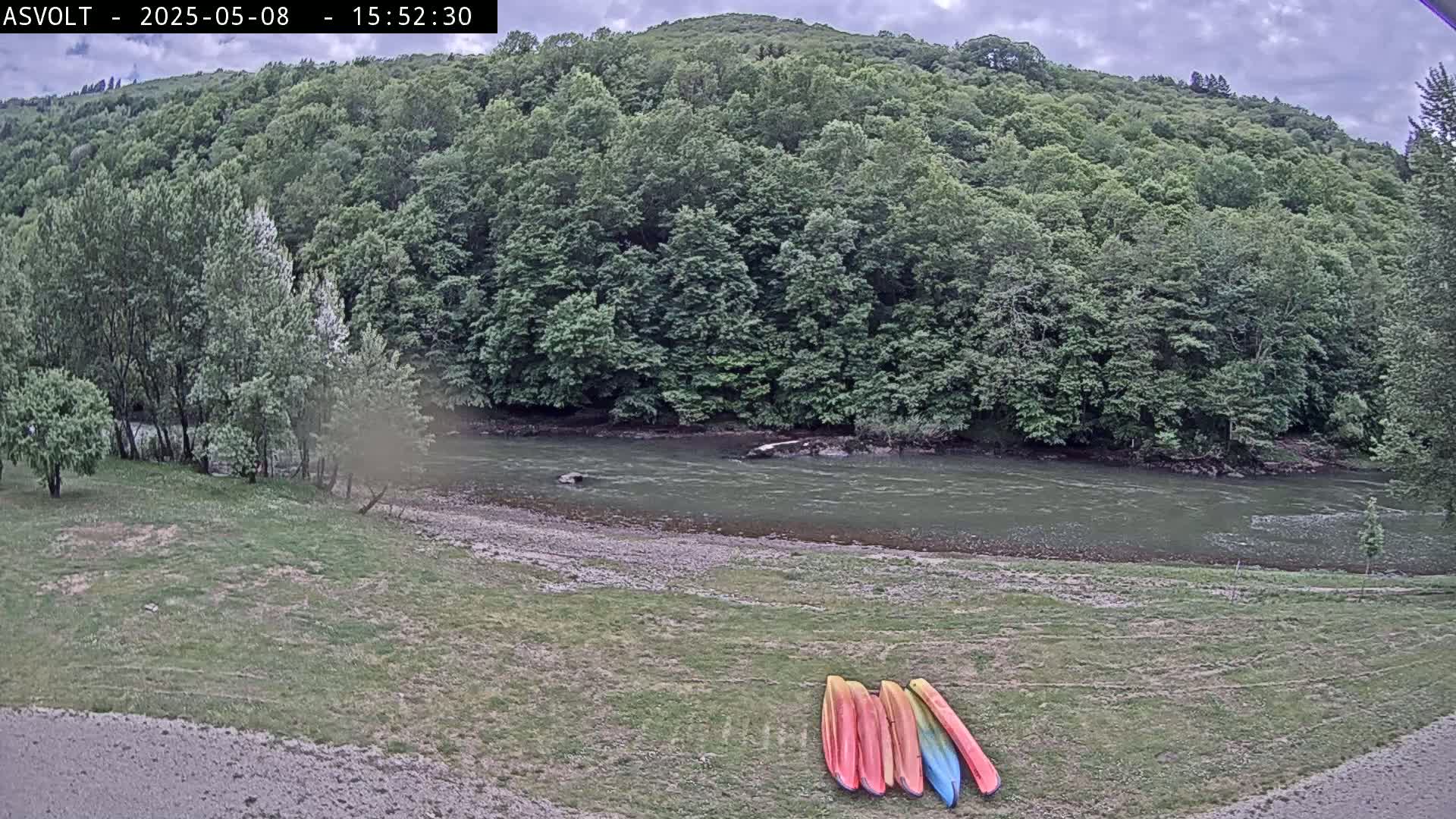 Several kayaks lie on a grassy riverbank next to a calm, grey river flowing past a lush green hillside under a cloudy sky.