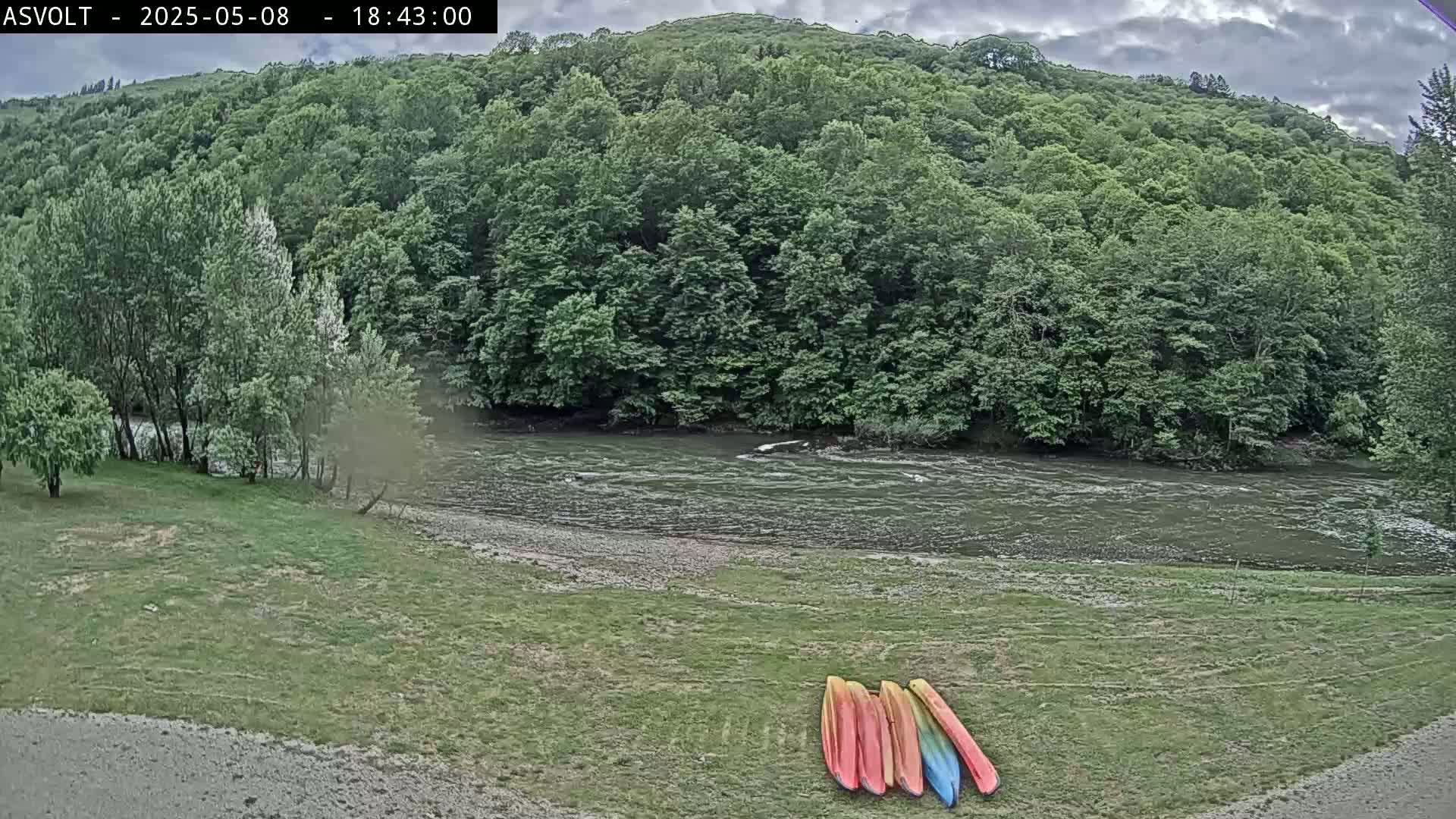 Several kayaks sit on a grassy riverbank next to a flowing river, under a partly cloudy sky, bordered by lush green trees and hills.