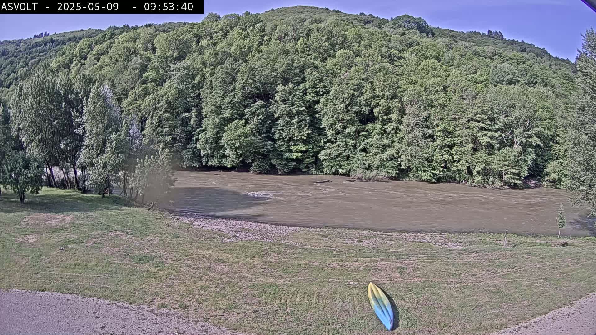 A muddy river flows beneath a lush green hillside on a sunny day, with a kayak resting on the grassy riverbank.