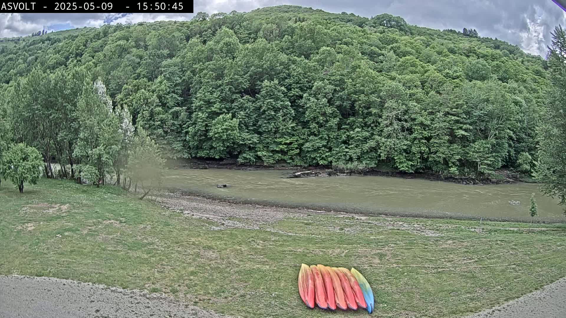 A muddy river flows beneath a lush green hillside under a cloudy sky, with several kayaks resting on a grassy bank.
