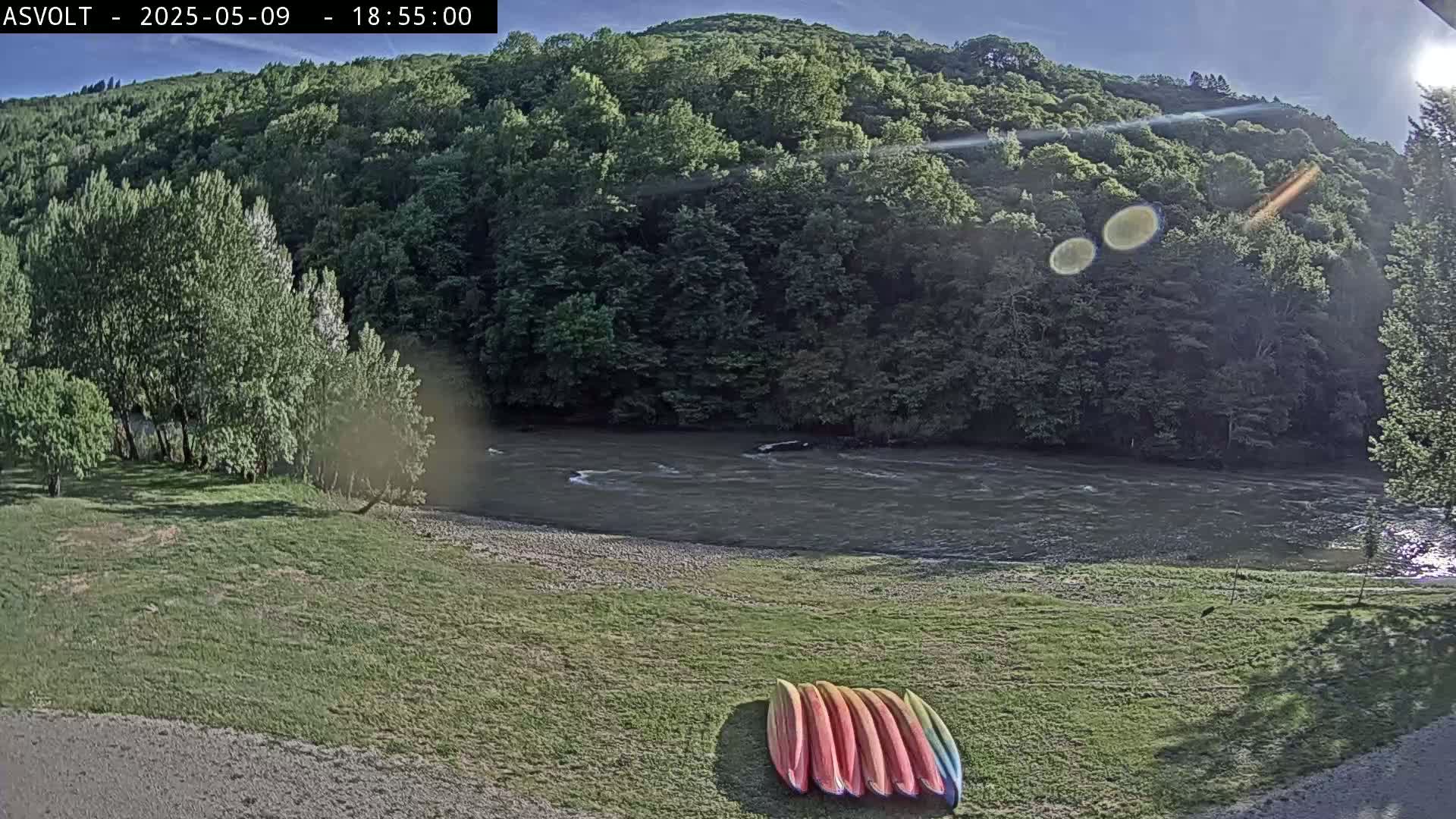 Several kayaks lay on a grassy bank beside a flowing river under a mostly sunny sky, backed by a lush green hillside.