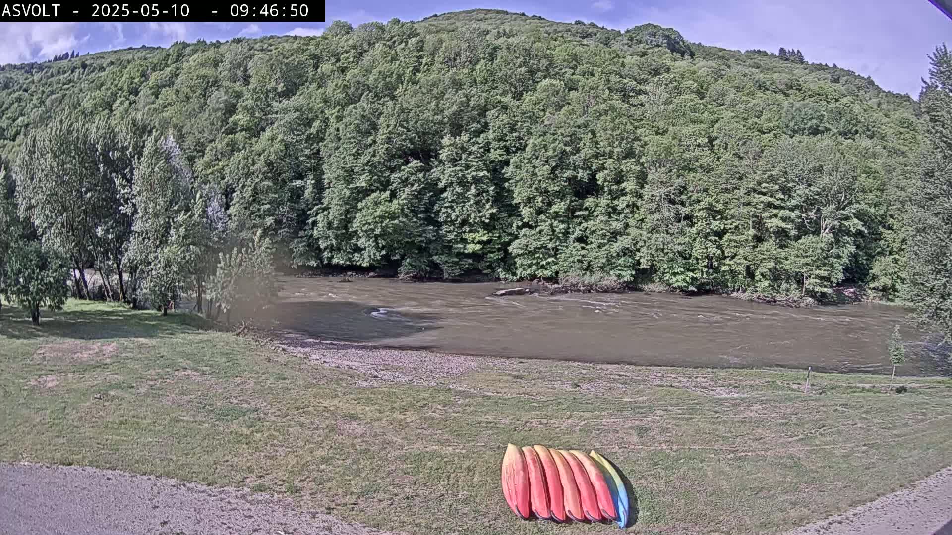 Kayaks are lined up on a grassy bank next to a muddy river flowing beneath a hill covered in lush green trees under a mostly sunny sky.